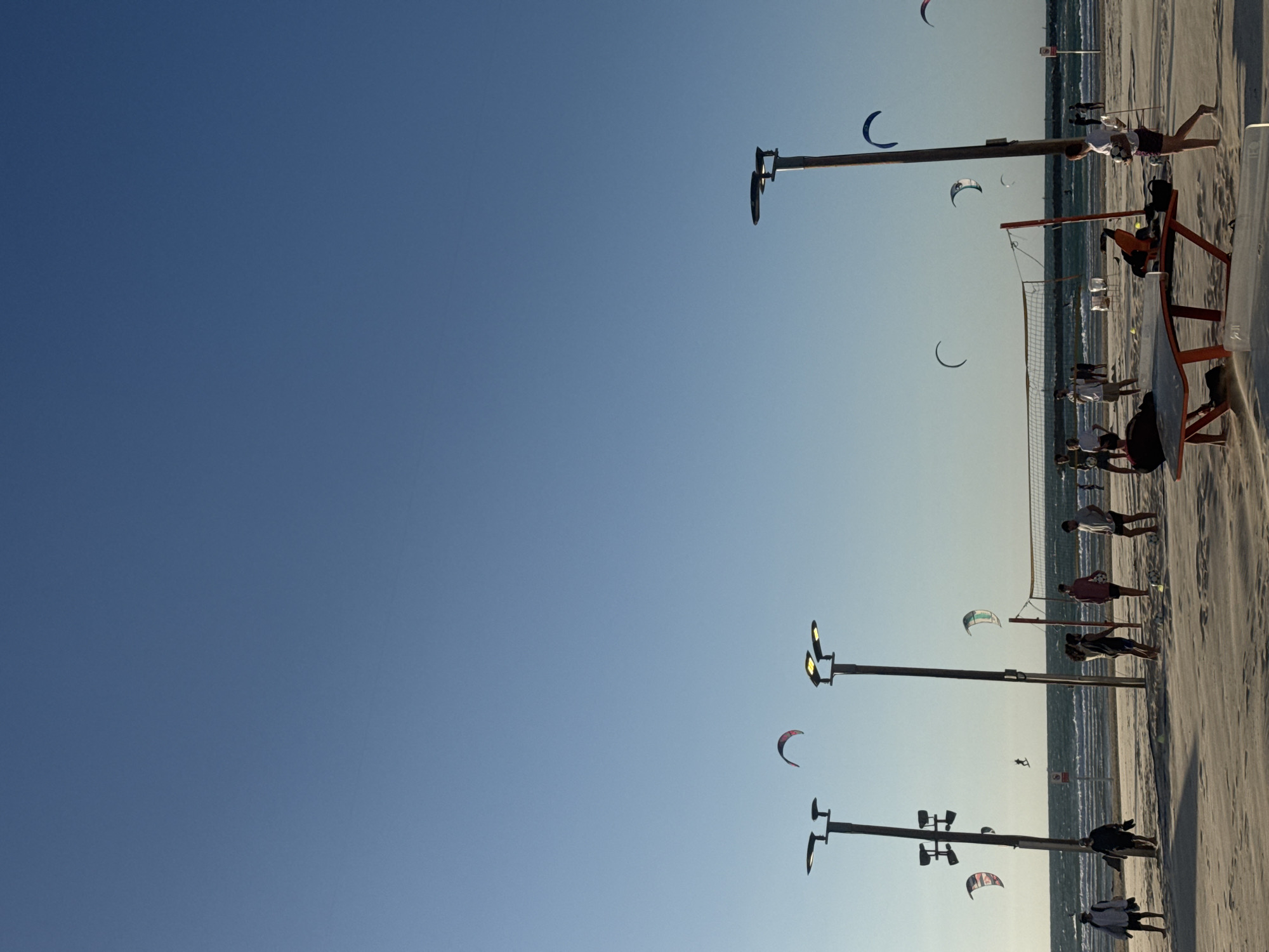 The image depicts a beach scene in Tel Aviv, Israel. The sky is a clear gradient of blue. On the right side of the image, the beach is visible with light sand. People are playing soccer and kitesurfing along the shoreline. There are lampposts spaced along the beach. A volleyball net is set up with people gathered around it. The sea stretches to the horizon.