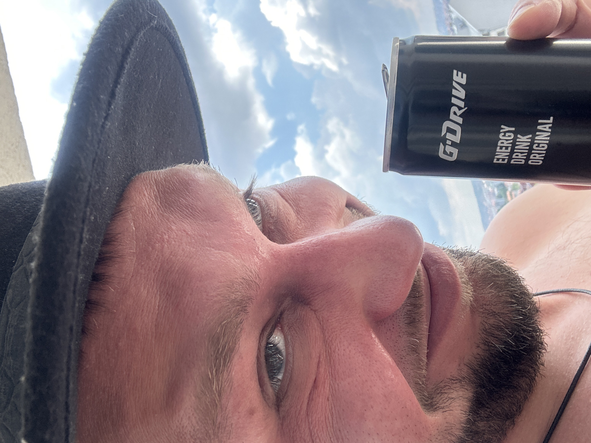 The media file is a close-up, upside-down selfie of a man holding a black can of "G-DRIVE ENERGY DRINK ORIGINAL." The man is wearing a dark hat and has stubble on his chin. His face is angled towards the camera, and his eyes are visible, looking slightly to the side. The background consists of a bright, cloudy sky, suggesting it is daytime and the weather is likely pleasant. The man's skin appears flushed, which could indicate he has been outdoors in the sun. The setting appears to be outdoors, possibly on a balcony or rooftop, given the view of the sky and distant buildings, consistent with a location in Zvezdara Urban Municipality, Serbia. The image conveys a casual, perhaps spontaneous moment captured outdoors.