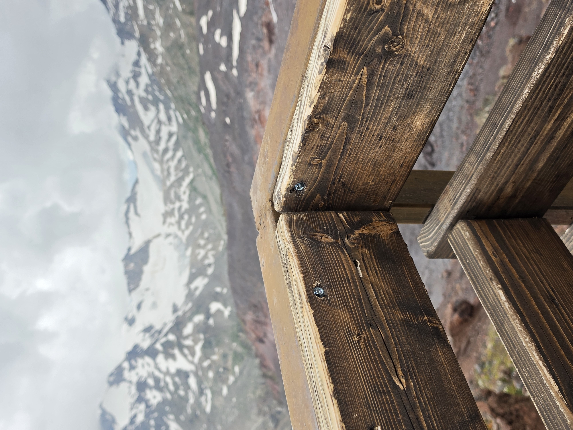 The image shows a close-up of a wooden railing in the foreground, with a mountain range in the background. The railing is made of dark, weathered wood and appears to be part of an observation deck or viewing platform. The mountains in the distance are covered in snow and clouds. The overall scene suggests a natural, rugged environment, possibly a hiking or scenic viewpoint. The city of Terskol, Russia, is known for its mountainous terrain and proximity to Mount Elbrus, a dormant volcano and the highest peak in Europe. This image aligns with the natural scenery one would expect to find in that region. There are no people or animals visible in the image.