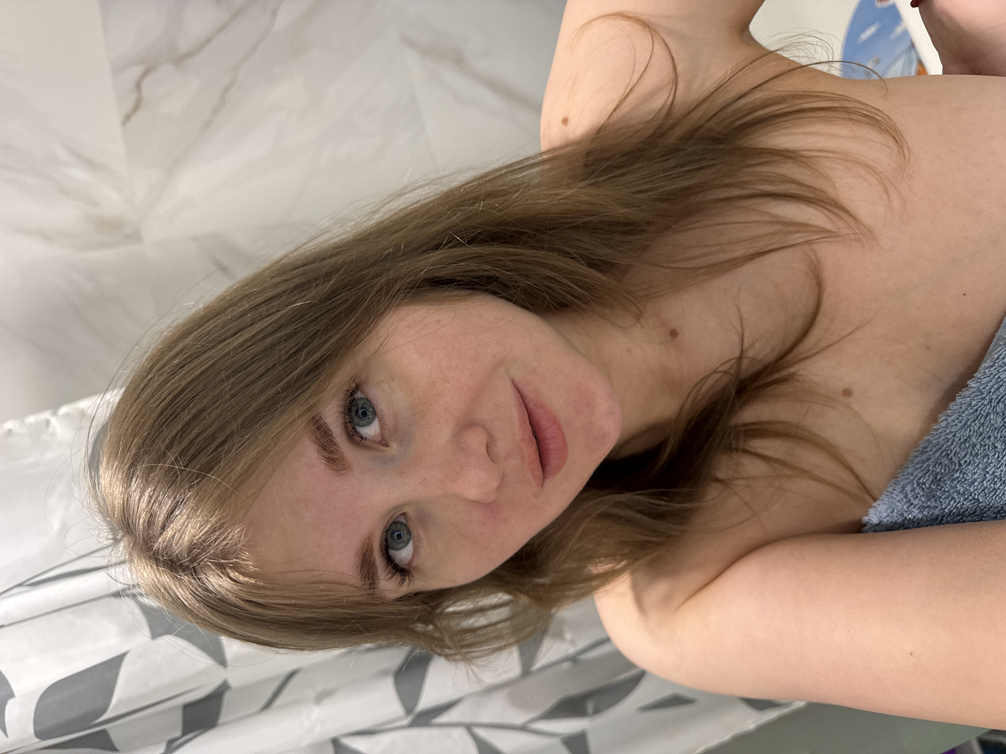 A woman with light brown hair and blue eyes is photographed from an elevated perspective. She is partially covered with a blue towel. The background includes a marble-patterned surface and a white shower curtain with a grey pattern. The city of Ферма, Russia, is not identifiable from the image.