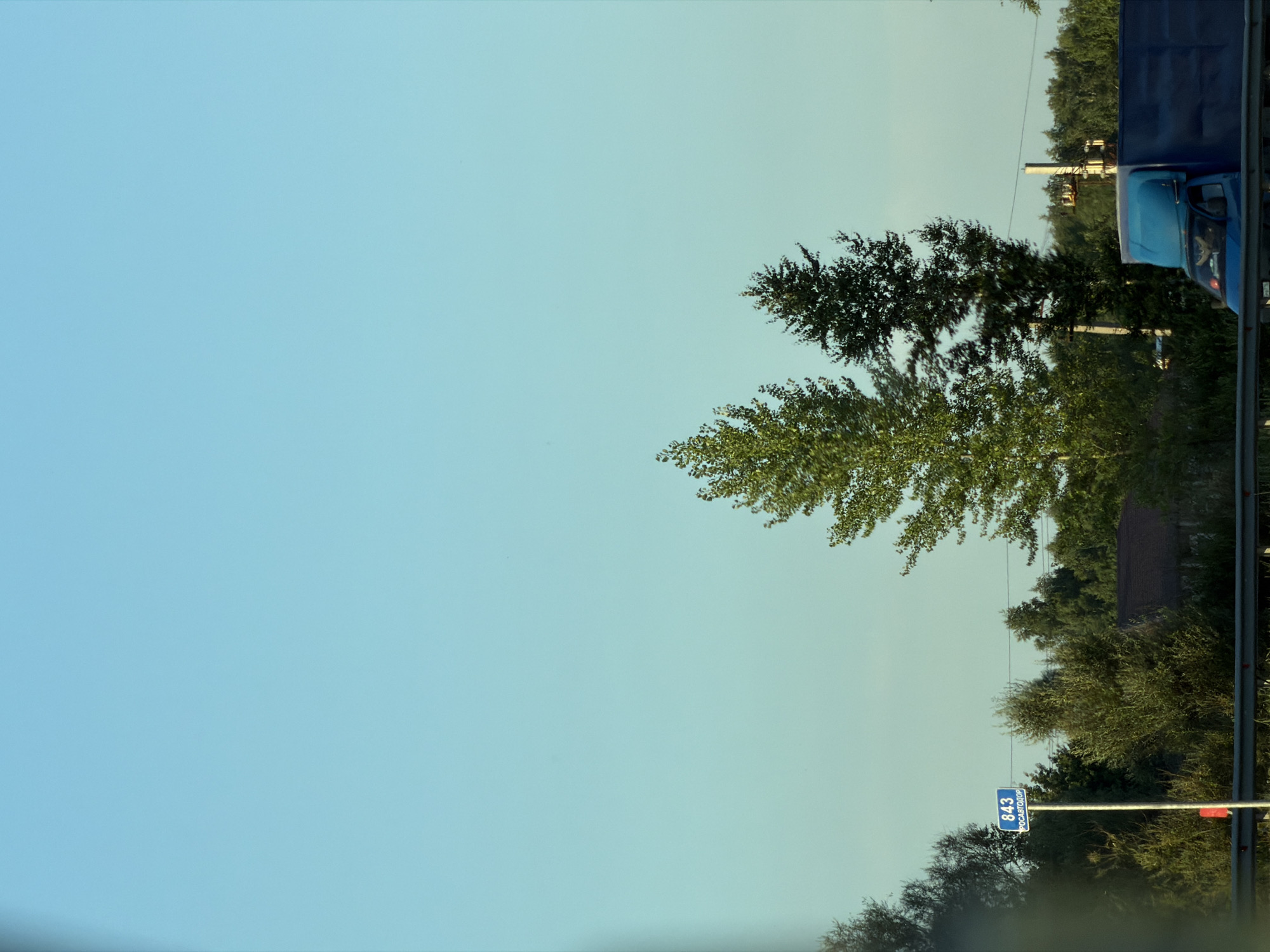 The image captures a landscape scene with a clear, pale blue sky dominating the upper portion. On the right side, a dark blue truck is partially visible, with its cab and part of the trailer showing. Trees with lush green foliage are scattered throughout the scene, with some taller trees reaching towards the sky. In the middle ground, there appears to be a building with a dark roof. Closer to the foreground, a blue street sign with white text is mounted on a pole. The text on the sign reads "843" and "РОСКАВТОДОР". The overall impression is of an outdoor setting, possibly a roadside view, with a clear sky suggesting a pleasant day. The light appears soft, indicating it might be early morning or late afternoon. The presence of the truck and the street sign suggests a location near a road, likely in a rural or suburban area.