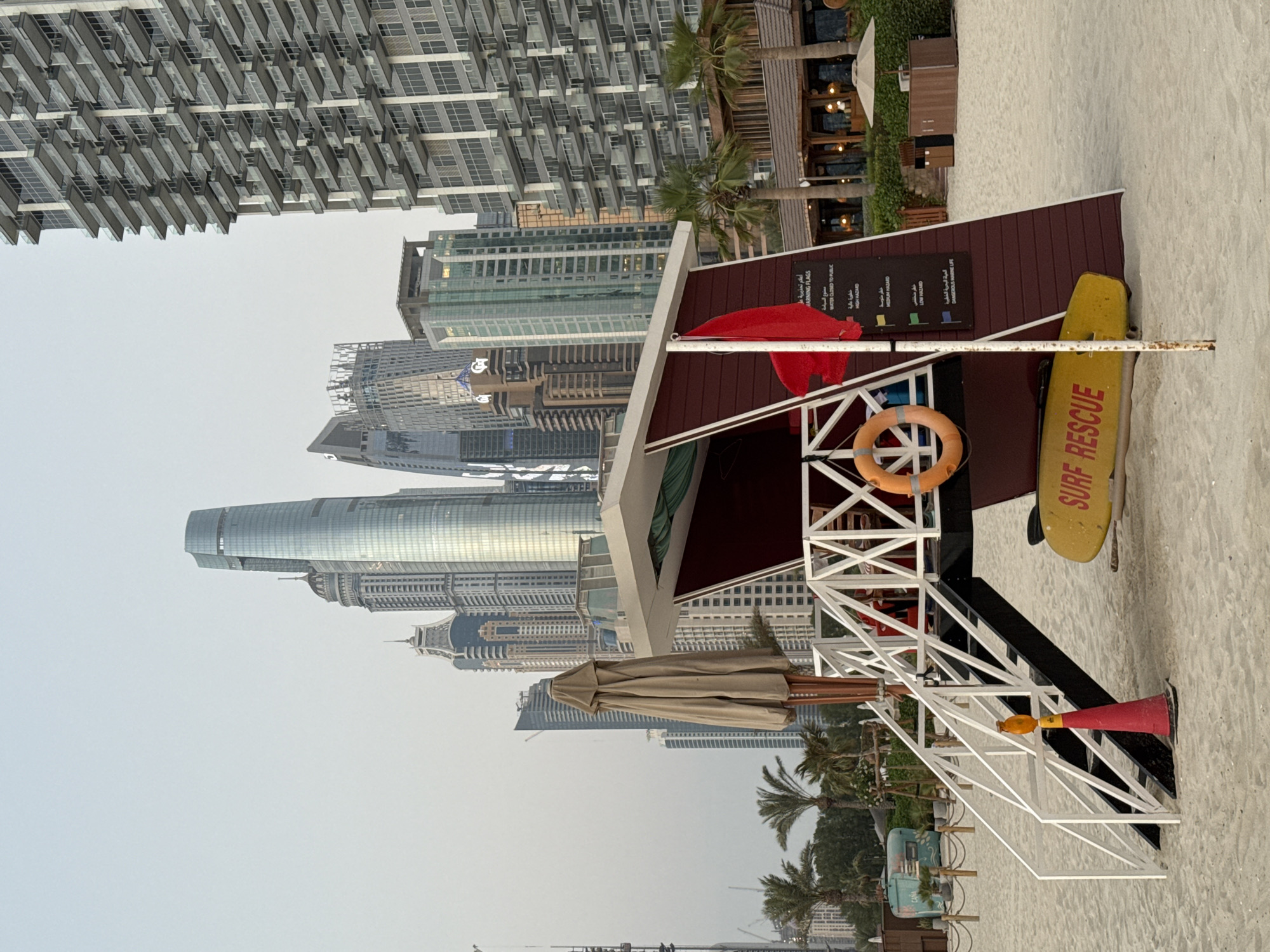 This image depicts a beach scene in Dubai, United Arab Emirates. The primary subjects are a lifeguard station, a yellow surfboard with "SURF RESCUE" written on it, and a life preserver. The lifeguard station itself is a multi-level structure with white railings and a maroon roof. There is a red flag on a pole near the station and a red and yellow horn at its base. In the background, several tall, modern buildings characteristic of Dubai's skyline are visible, along with palm trees and beach umbrellas. The sky is overcast, suggesting a hazy or cloudy day, and the lighting indicates it is daytime. The beach is covered in sand. Visible text includes English and Arabic warning flags and hazard information related to water safety, such as "HIGH HAZARD," "MEDIUM HAZARD," "LOW HAZARD," and "DANGEROUS MARINE LIFE." The scene appears to be set up for beachgoers, with the lifeguard station ready for use. There are no people or discernible activity taking place in the frame, giving a sense of stillness to the scene.