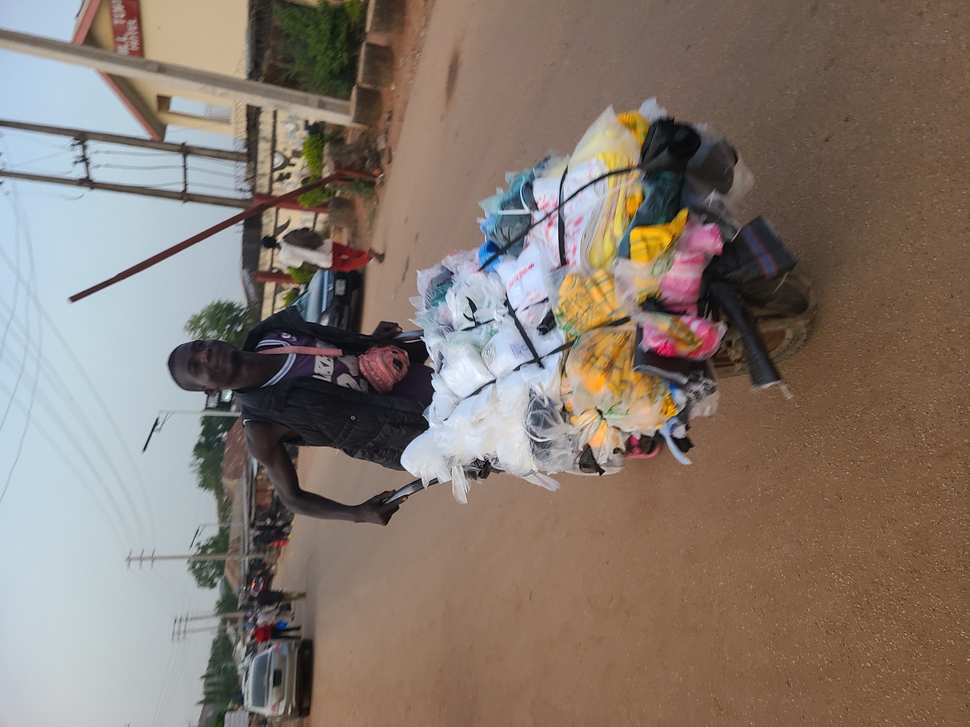 The image shows a man walking along a paved road in Jos, Nigeria, pulling a cart overloaded with bundles of plastic bags and other items. The bags are of various colors, including white, yellow, pink, and black, and are secured with black straps. The man is wearing a black sleeveless vest and a jersey. The road appears to be a residential or commercial street with buildings and vehicles visible in the background. There are other people walking on the road and some parked cars. The lighting indicates it's either morning or late afternoon. Electrical poles and wires are visible along the road.