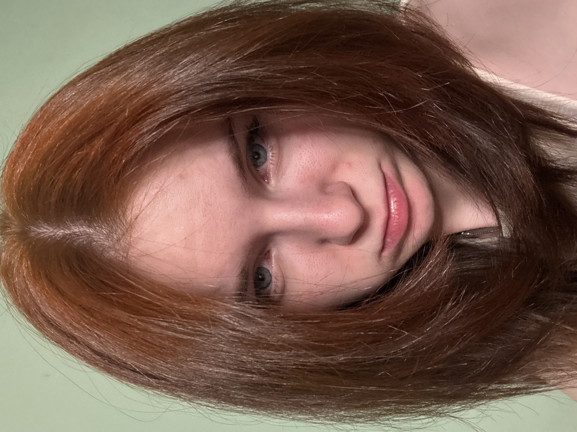 A close-up, slightly rotated image of a person with red hair. The person has fair skin and light-colored eyes. The background appears to be a light green or pale color. The scene's location is Unknown, Unknown.