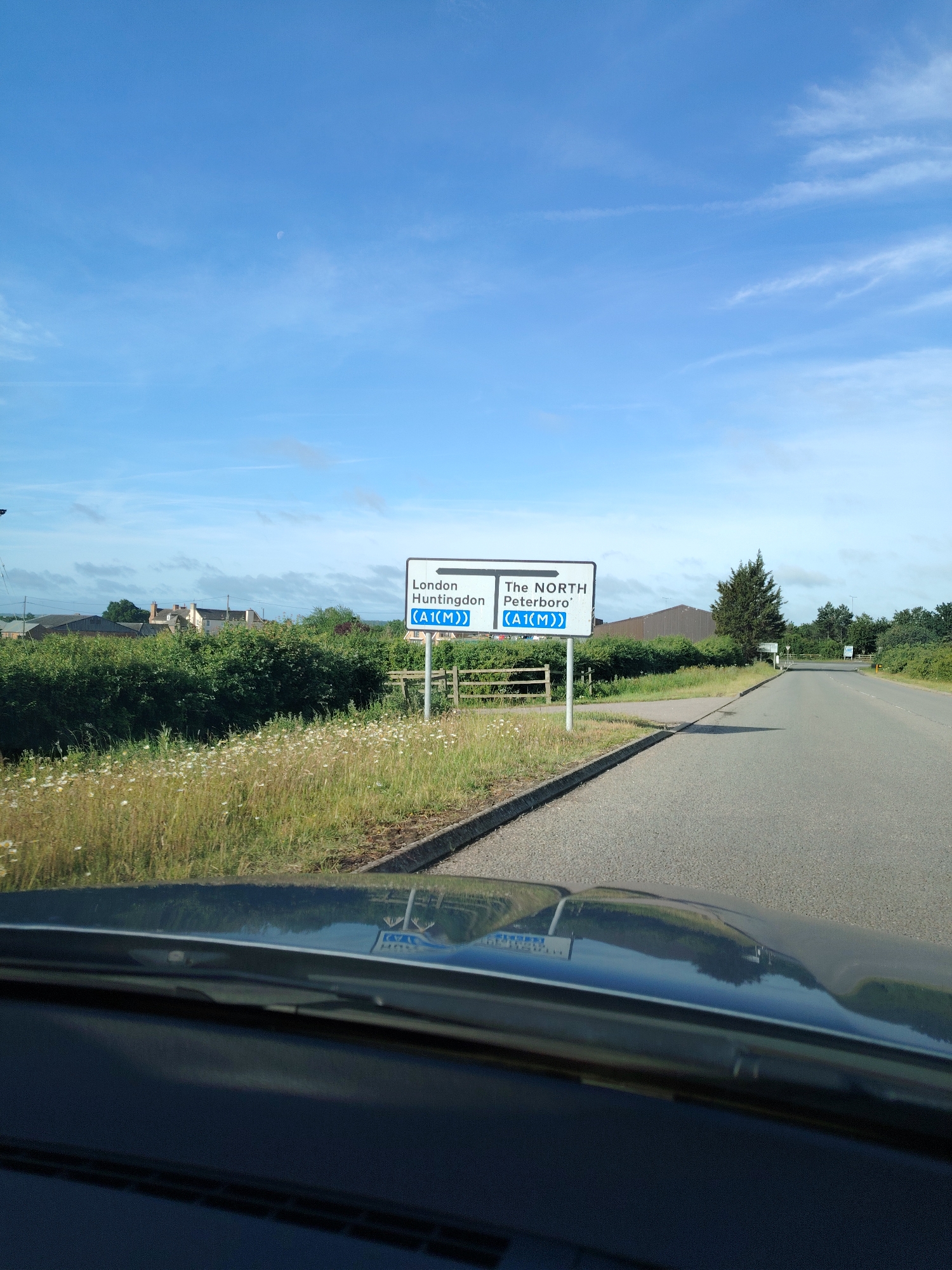The image depicts an outdoor scene on a clear day, captured from within a vehicle, evidenced by the dark dashboard and reflections visible in the foreground. The sky, occupying the upper half of the frame, is a clear light blue with scattered wispy white clouds, and a faint crescent moon is discernible in the upper-middle section.
A paved asphalt road curves gently to the right, extending into the mid-distance. A concrete curb separates the road from a grassy verge on the left. This verge is overgrown with green and dried grasses, densely populated with small white flowers resembling daisies. Behind the verge, a continuous line of dense green bushes forms a hedge.
In the mid-ground, two prominent white road signs with blue borders are positioned roadside on metal posts. The left sign features a left-pointing arrow and reads "London Huntingdon (A1(M))". The right sign displays a right-pointing arrow and reads "The NORTH Peterboro' (A1(M))". Behind the hedges to the left, several light-colored buildings with dark roofs are visible. Further to the right, behind the second sign, a large, dark, industrial-style building with a pitched roof stands amongst trees.
The presence of road signs indicating "Huntingdon," "London," "Peterboro'," and the "A1(M)" precisely places this location at an access point to the A1(M) motorway, near Huntingdon, within Huntingdonshire, United Kingdom. The signs direct traffic south towards London and Huntingdon, and north towards Peterborough and "The NORTH," indicating a junction or interchange along the A1(M).