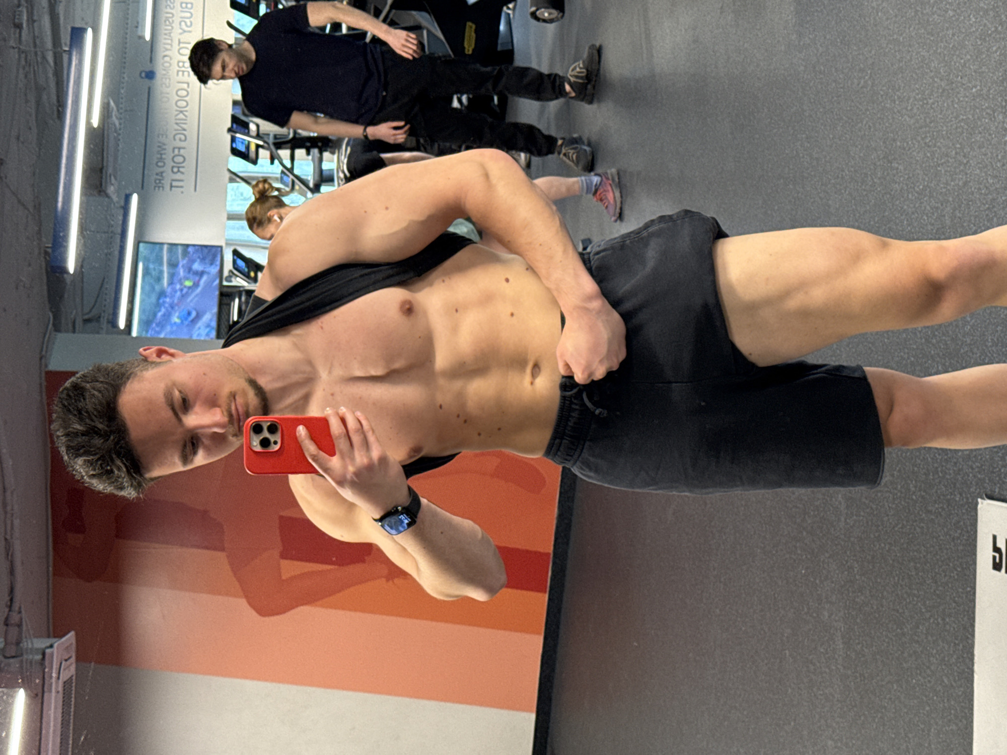 A young adult male takes a shirtless selfie in a gym, reflected in a mirror. The image is rotated 90 degrees clockwise. The main subject has short dark hair and a beard, showcasing a muscular torso with visible abs and freckles/moles. He wears black shorts and a black smartwatch on his left wrist, holding a red iPhone in his left hand to capture the image. His expression is neutral as he looks into the phone's camera.
In the background, other gym patrons are engaged in activities. A second young adult male, also with dark hair, wears a dark t-shirt and black pants while standing near cardio equipment. Partially visible behind him is another individual, likely female, with hair tied in a bun, wearing a light-colored top and pink athletic shoes, also using gym machinery.
The setting is a well-lit fitness center in Sofia, Bulgaria, indicated by bright, rectangular ceiling lights. The floor is dark gray, and a large, abstract wall graphic in shades of orange and red is reflected in the mirror behind the main subject. Visible text on a mirrored sign in the background reads, "YOU'RE SO BUSY TO BE LOOKING FOR IT!". The overall scene suggests a routine workout environment.