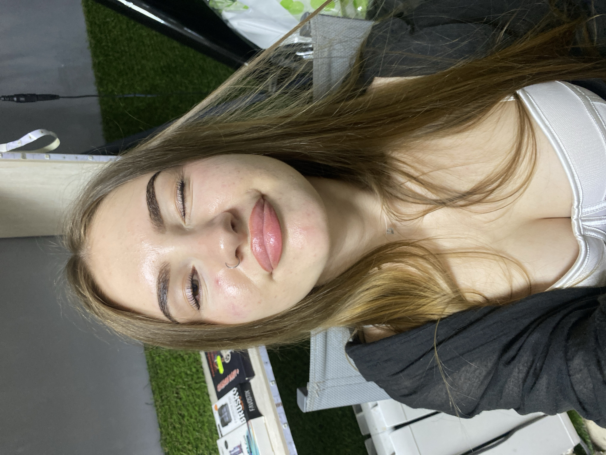 A young woman with light brown hair is looking at the camera and winking. She is wearing a black open cardigan over a white top. The woman has a ring in her nose and is wearing light makeup. She has a pouty expression and her lips are glossy. In the background, there is a wall with artificial grass and some shelves with products. The scene appears to be indoors. There is no information to suggest the location is in Dyakovo, Russia.