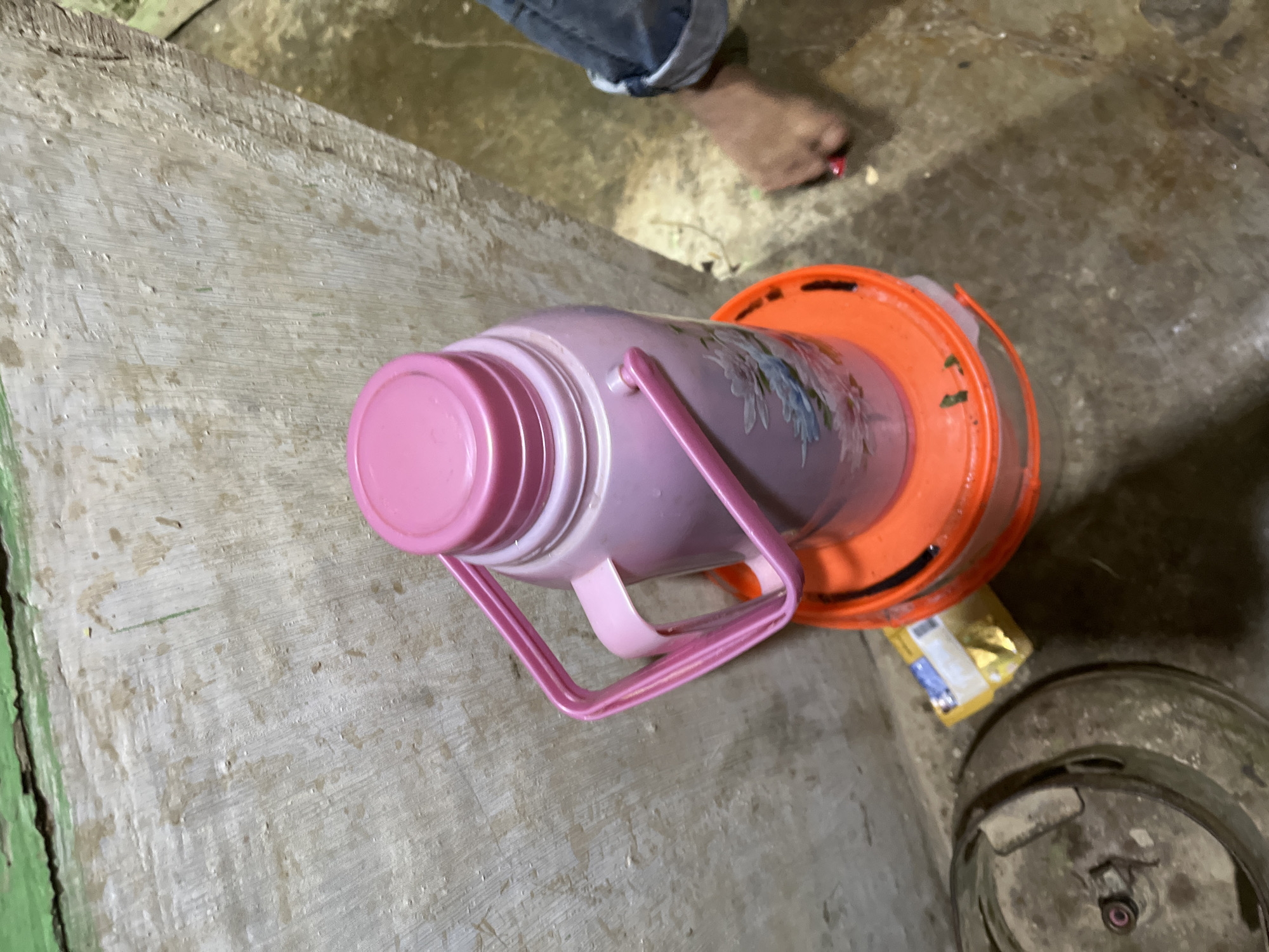 The image shows a pink water jug with a floral pattern placed on a rough, light-colored surface, possibly wood. The jug has a pink handle and lid. It is sitting inside an orange plastic container. To the right of the jug, there's a small, yellow, foil-wrapped packet on the floor, which appears to be food packaging. Further to the right, a round, metallic object with visible screws and nuts is partially visible. In the background, a person's legs and feet are visible. The person is wearing blue jeans that are rolled up at the ankles, and one foot is bare, showing red nail polish. The scene appears to be indoors, with a concrete or tiled floor. There are no obvious indicators of time of day or weather. The overall impression is of an everyday domestic or utility setting.
