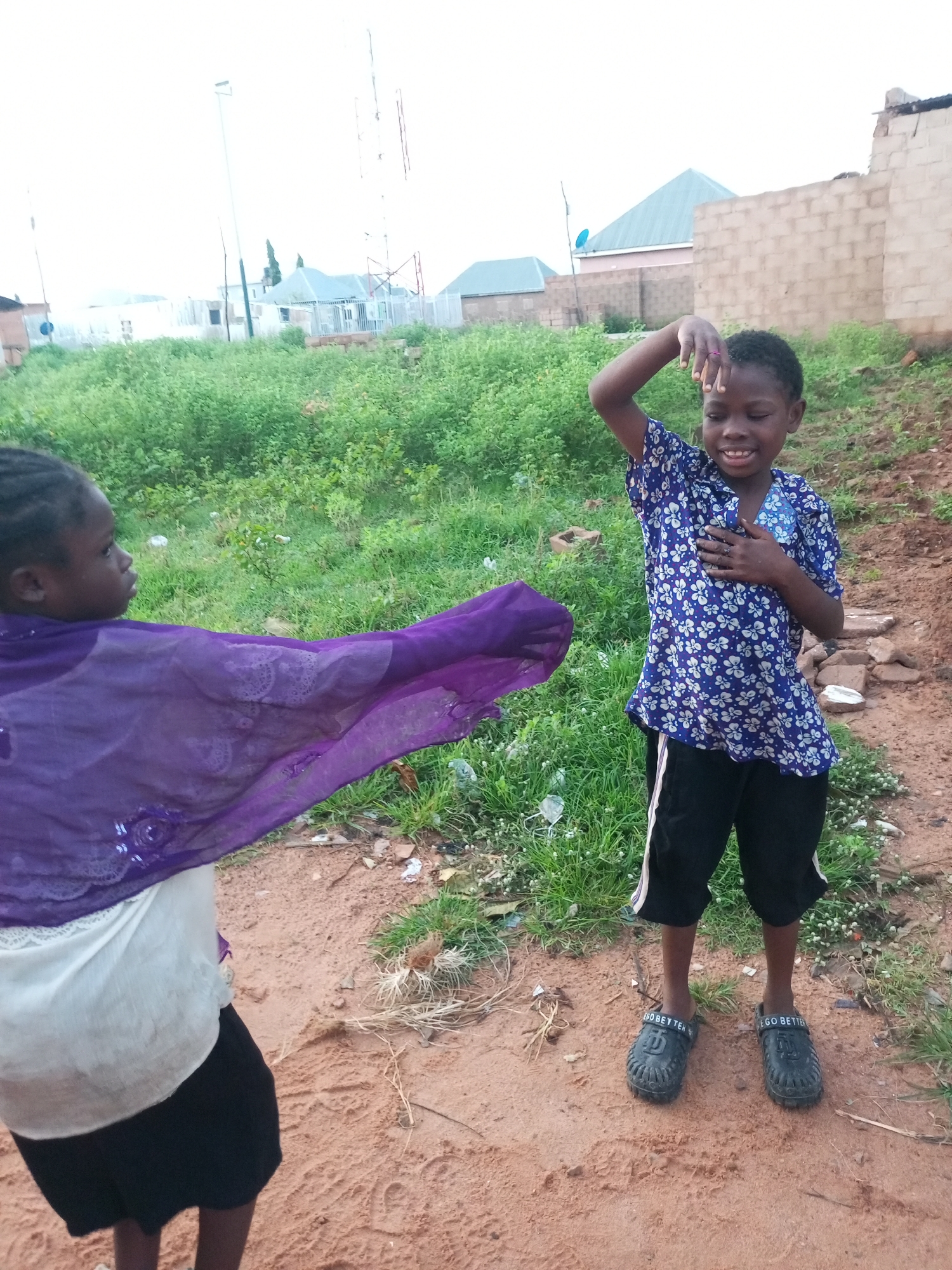 In Tempure, Nigeria, two young girls are interacting outdoors. The girl on the right is wearing a blue and white floral print shirt and black shorts. She has her right arm raised, with her hand near her forehead, and her left hand is placed on her chest. She is smiling. The girl on the left is wearing a white top and a black skirt, with a vibrant purple scarf draped over her arm. The background features green vegetation, some scattered debris, and buildings with light-colored walls and blue roofs. A tall communication tower is visible in the distance. The ground is composed of reddish-brown soil and sand.