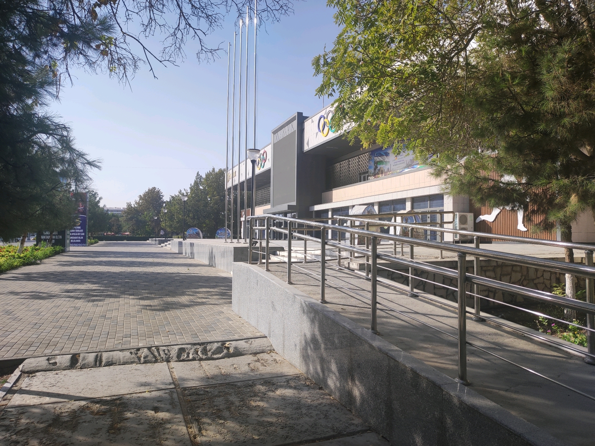 The image shows an outdoor scene in Navoiy City, Uzbekistan, featuring a wide paved walkway leading towards a large modern building with Olympic rings prominently displayed on its facade. The building is light-colored with several windows and appears to be an event or sports venue. In front of the building, there are tall flagpole structures. A metal railing lines a ramp or pathway that curves to the right, with shadows from trees cast across the paved area. The environment suggests a public space with vegetation including trees and some greenery along the sides. The sky is clear and blue.