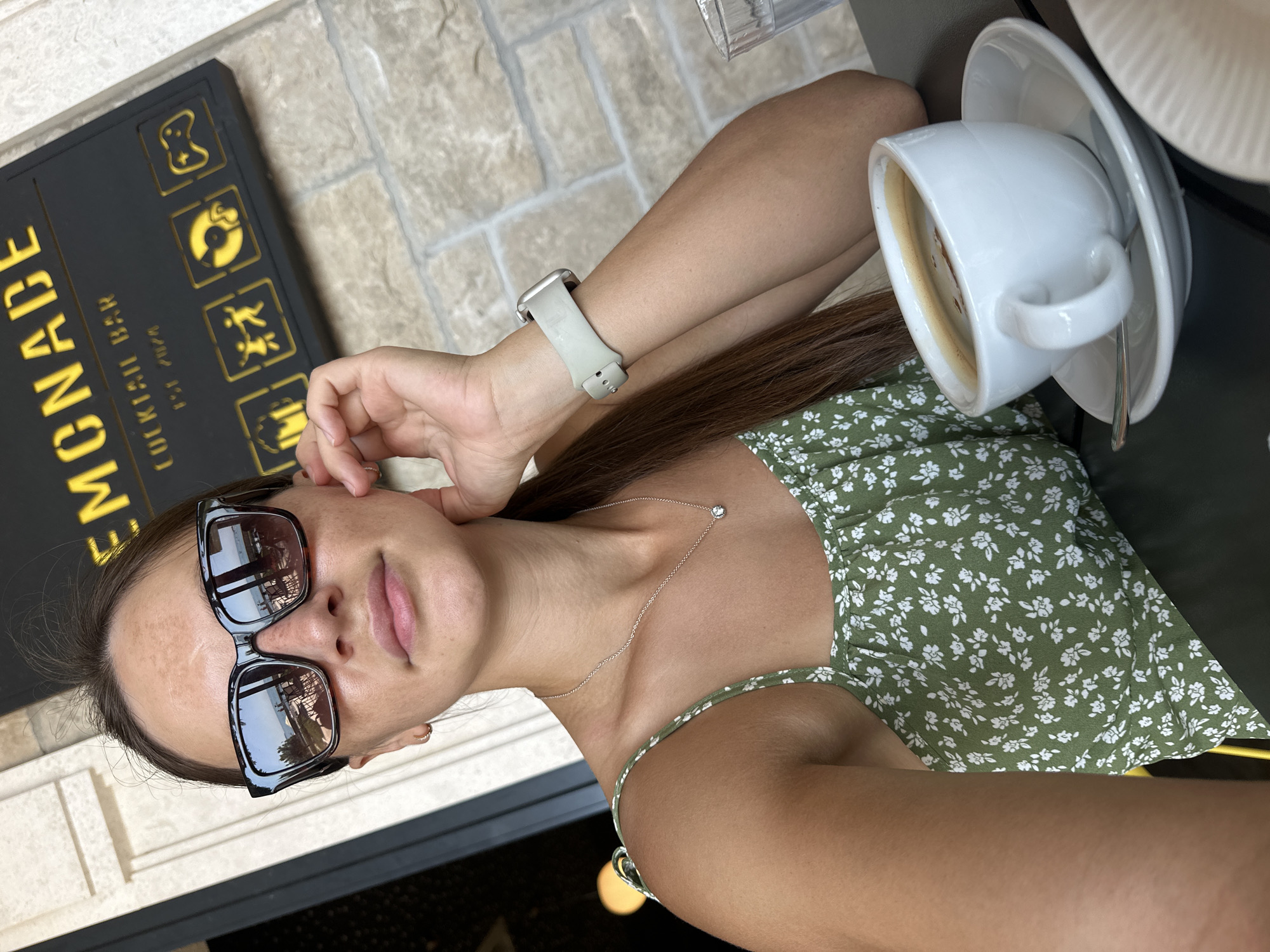 A woman with sunglasses and a green floral top is taking a selfie with a cup of coffee. She is sitting at a table with a sign that reads "LEMONADE COCKTAIL BAR EST. 2024". The background appears to be an outdoor seating area with a stone wall and a building. The woman is resting her hand on her chin and looking at the camera. The coffee is in a white cup with a saucer.
