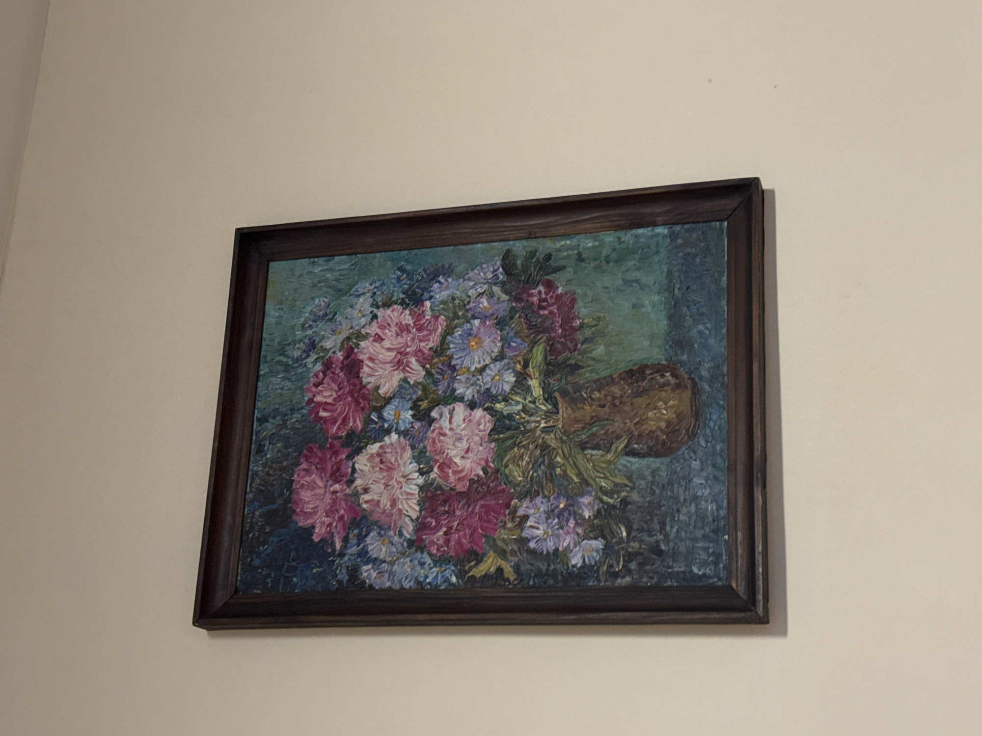 A framed painting of flowers is hanging on a light-colored wall. The painting depicts a vase full of colorful flowers. The vase is brown and squat, and the flowers are various shades of pink, purple, and blue. The background of the painting is a mottled blue and green. The painting is framed in a dark brown wooden frame. The location of the image is not discernible as Sofia, Bulgaria.
