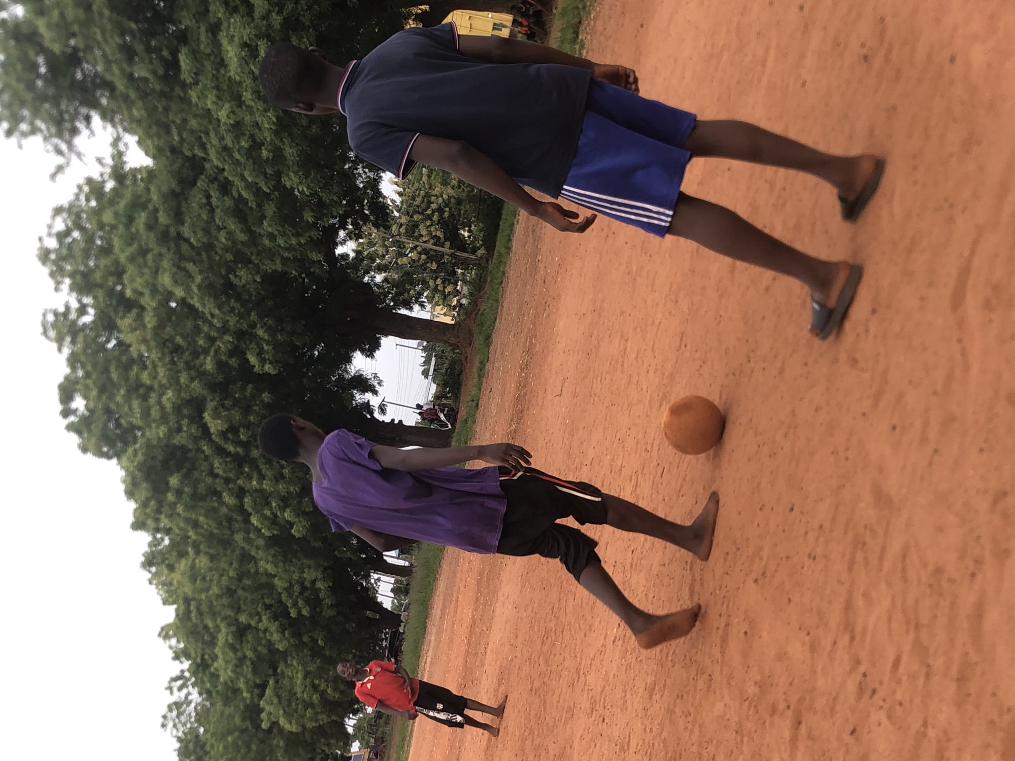 This image depicts three young males playing football (soccer) on a reddish-brown dirt field in an outdoor setting in Nsugbe, Nigeria. The sky is bright and appears overcast or indicates early morning/late afternoon, as there are no harsh shadows.
In the foreground, a young person wearing a purple t-shirt and black shorts is positioned barefoot, seemingly about to engage with a light brown football resting on the ground. Directly opposite, in the mid-ground, another individual in a dark blue polo shirt and blue shorts, wearing flip-flops, stands observing the play. Further in the background, to the left, a third, smaller person wearing a red t-shirt and black shorts, also barefoot, is visible, smiling towards the camera.
The scene is surrounded by numerous large, leafy green trees, suggesting a natural or semi-urban environment. In the distant background, faint structures, including a yellow container or building and utility poles with lines, can be discerned. The informal game, played barefoot on a dirt surface, highlights a casual and recreational activity. No text is visible in the image.
