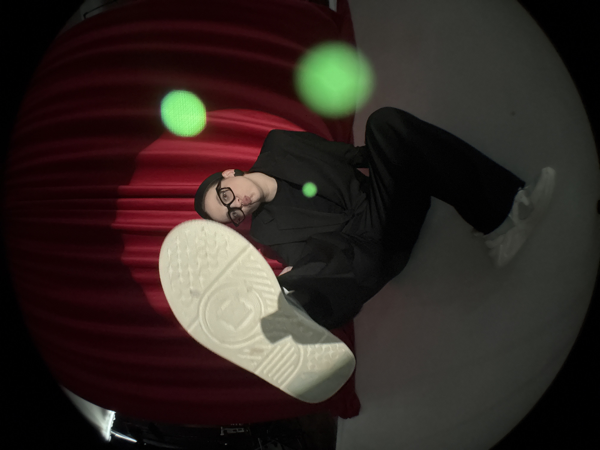 A person wearing a black suit, glasses, and a black beanie lies diagonally across a red and white background. The person's left leg is extended towards the camera, showing the sole of a white sneaker. There are three glowing green orbs in the air. The image has a fisheye lens effect with a dark, circular border. The setting appears to be a studio or staged environment, not a real-world location in Chaikovsky, Russia, based on the provided information.