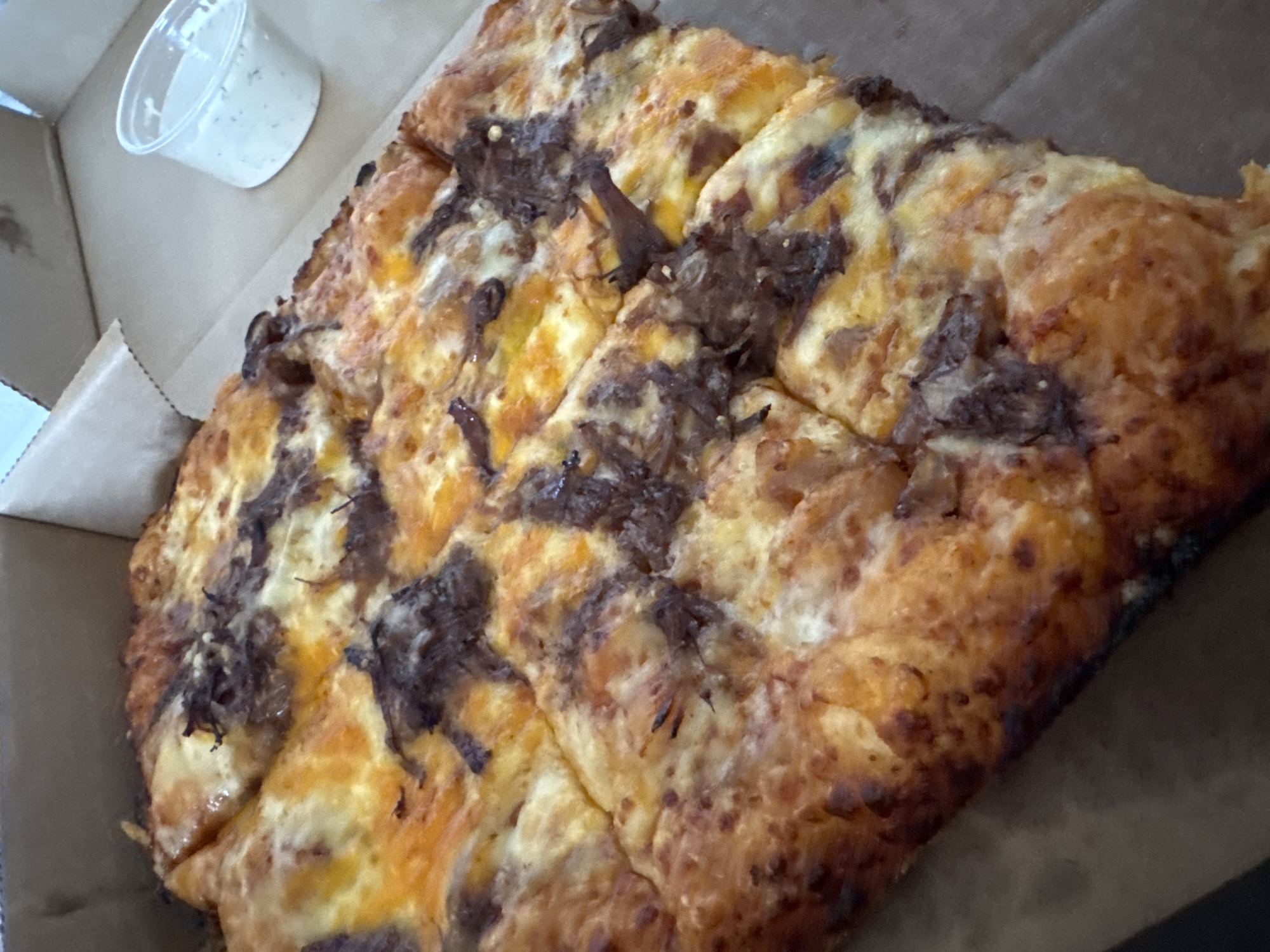 A cardboard pizza box contains a square pizza, cut into irregularly sized pieces. The pizza appears to have a golden brown crust and is topped with cheese and dark brown, possibly caramelized, onions. A small, clear plastic container filled with a white sauce (likely ranch dressing or similar) sits in the corner of the box. The scene is indoors, likely at a restaurant or in a home. There is no indication of this scene being located in Sofia, Bulgaria.