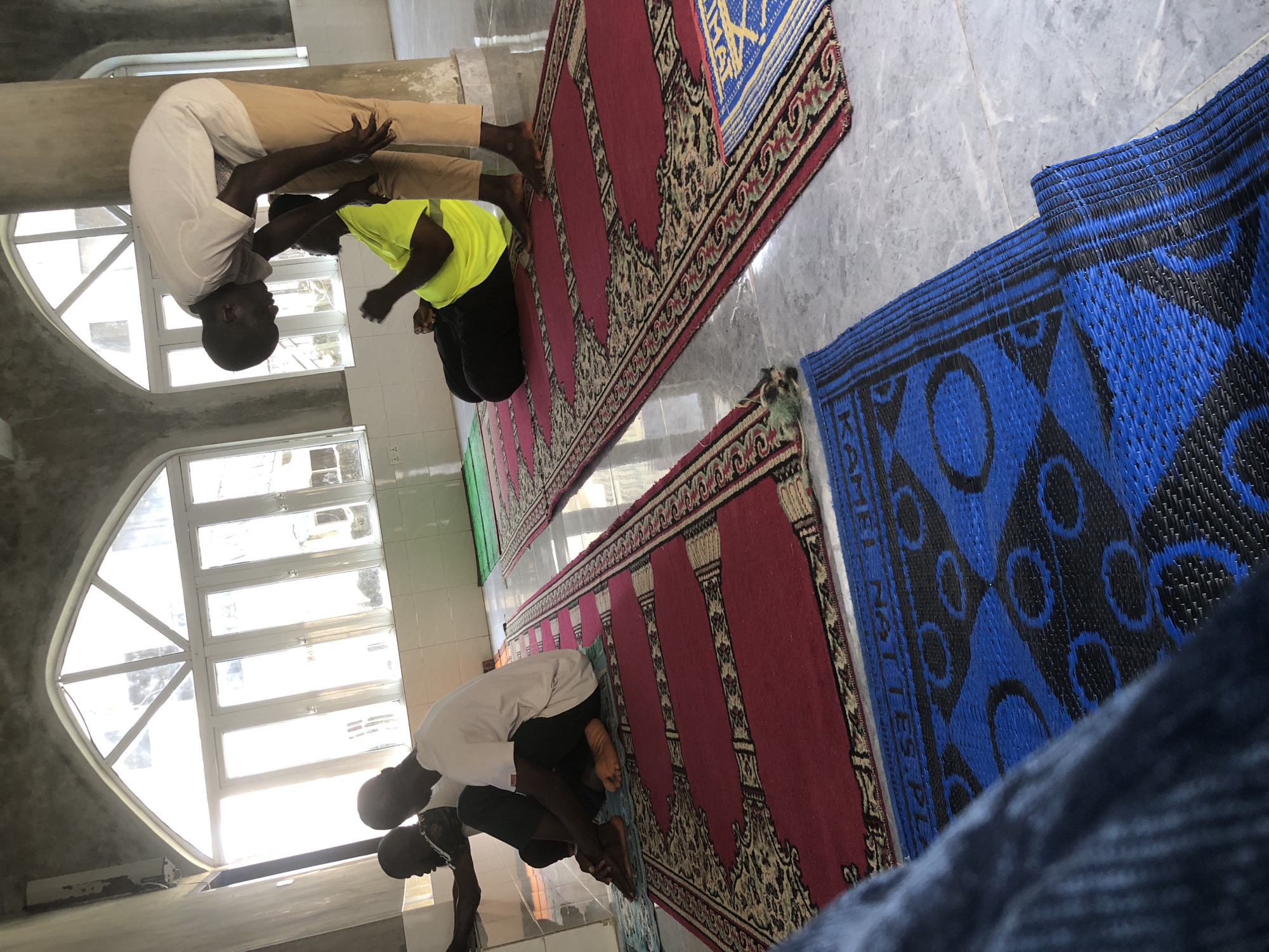 In the city of Unknown, Unknown, three individuals are depicted indoors, appearing to be engaged in prayer within a mosque or prayer hall. Sunlight streams through a large, arched window with multiple panes, illuminating the space and casting light on the tiled floor. Two men are seated on prayer mats, one in a white t-shirt and dark pants, and another in a bright yellow t-shirt and dark pants. A third man, standing and wearing a white t-shirt and beige pants, is positioned behind the seated individuals, leaning over them. The room features arched architectural elements and the floor is covered with several prayer mats of various colors and patterns, including red, blue, and green. The walls are a neutral color, with some tiled sections visible.