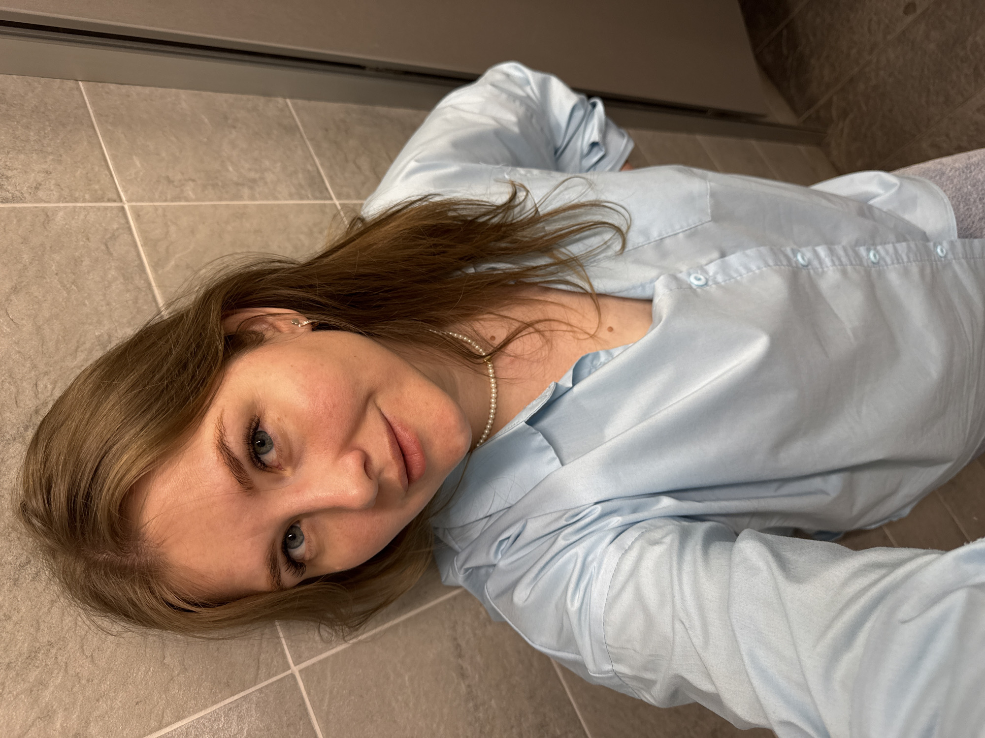 A young woman with light brown hair and blue eyes lies on a gray tiled floor. She is wearing a light blue button-down shirt and a pearl necklace. The floor is likely in a bathroom. The image does not provide information to confirm the location in the city of Perm, Russia.