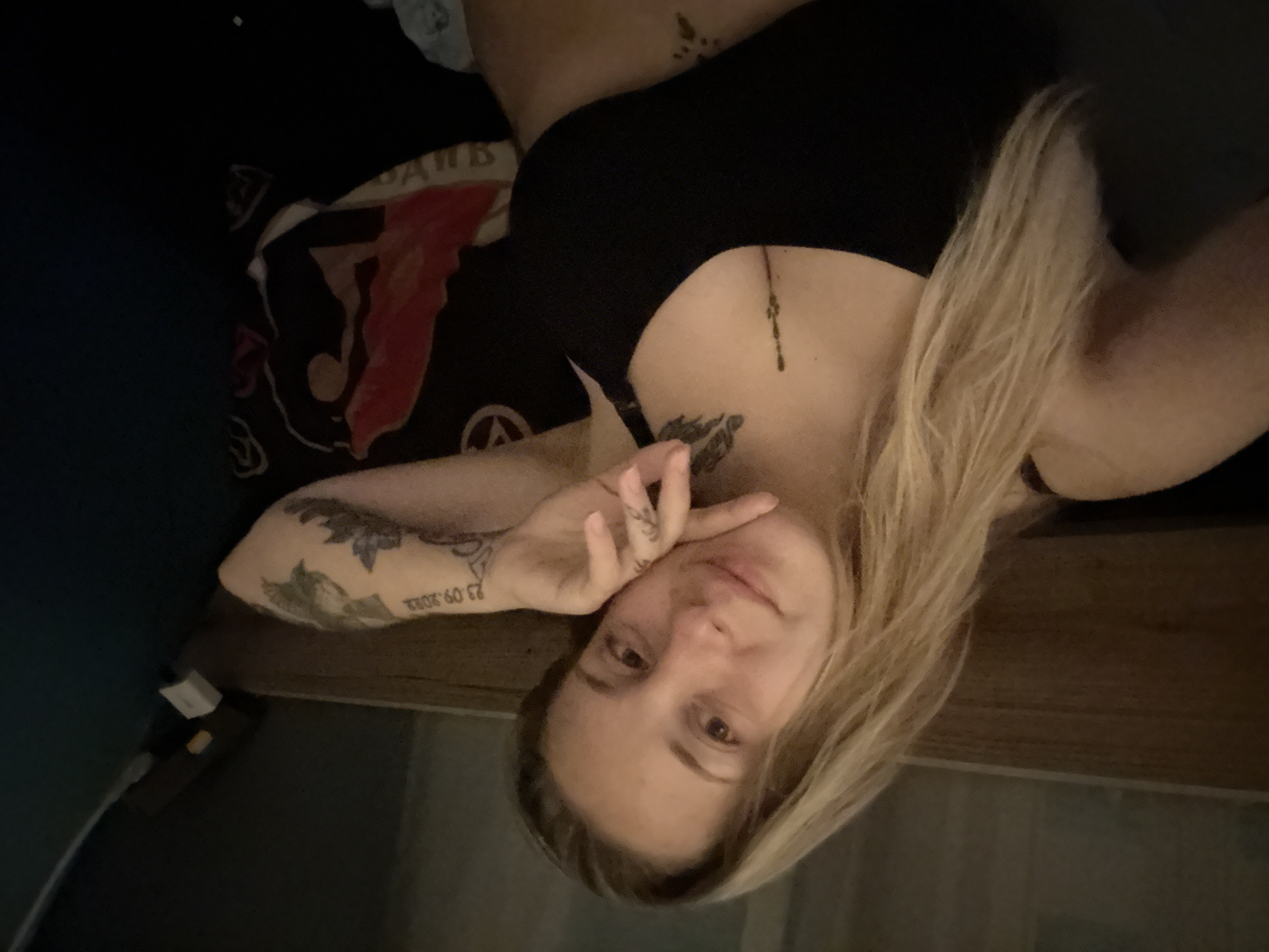 The image shows a woman with blonde hair wearing a black top. She has tattoos on her arm and chest. She is lying down, possibly in bed, with her head resting on her hand. The background appears to be a bedroom with a wooden headboard and dark colored bedding. There is a pillow with a design on it behind her. The scene is dimly lit, suggesting it might be nighttime or indoors with low lighting. There is no visible indication of the location being in Plovdiv, Bulgaria, or any urban setting.