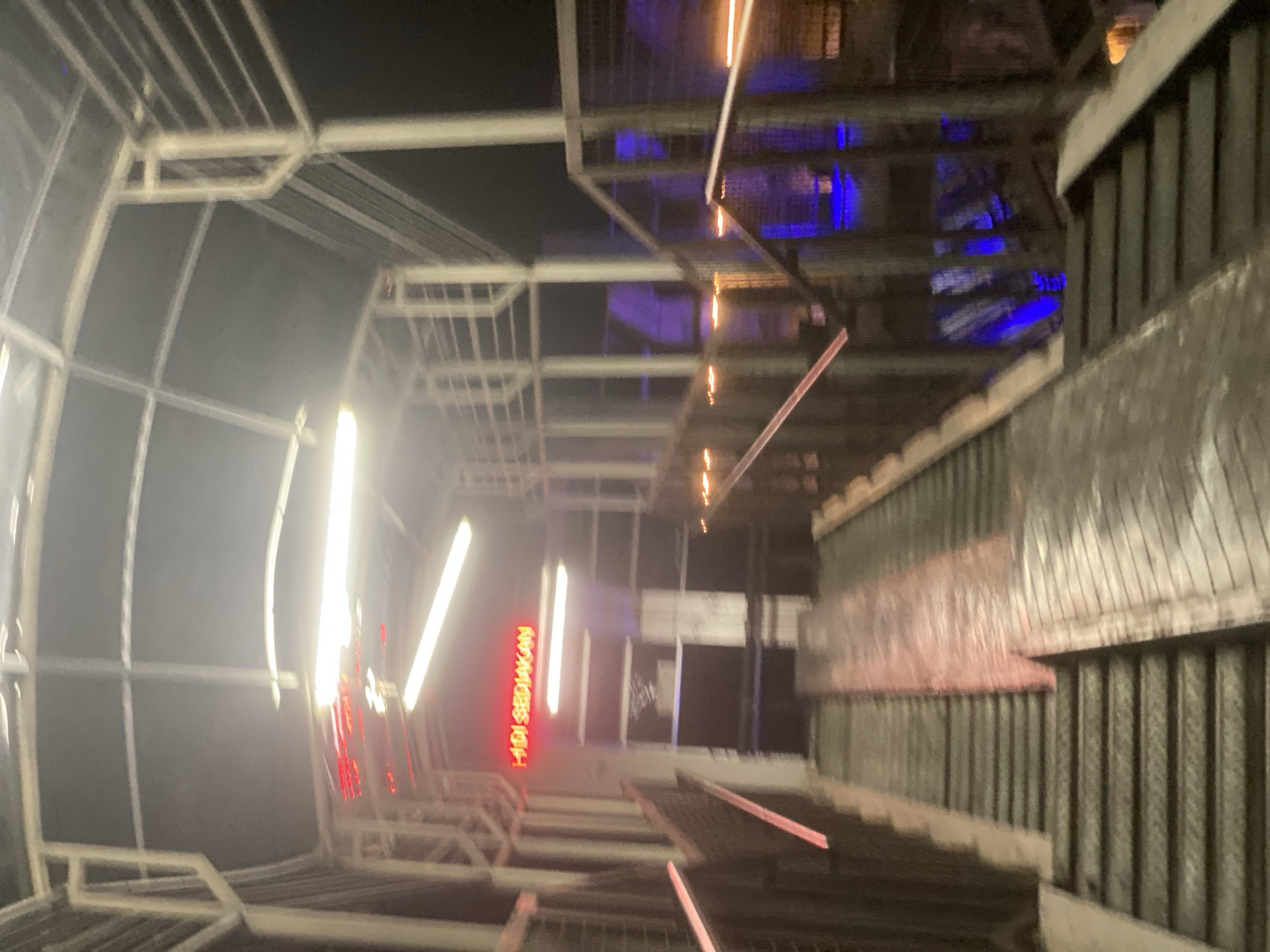This is a blurry, low-light photograph of an elevated walkway or pedestrian bridge at night. The structure appears to be made of metal, with a covered roof supported by white beams. Red neon lights are visible on the left side of the structure, spelling out "HADI SEDIAKAN" in Indonesian. The walkway is equipped with both stairs and a ramp, suggesting accessibility. Metal railings line the sides of the walkway. In the background, there are glimpses of blue and orange lights, possibly indicating other areas or businesses. The overall impression is that of a somewhat industrial or utilitarian urban environment, potentially a transportation hub or an elevated city pathway. The scene is located in Banjarbaru, Indonesia.