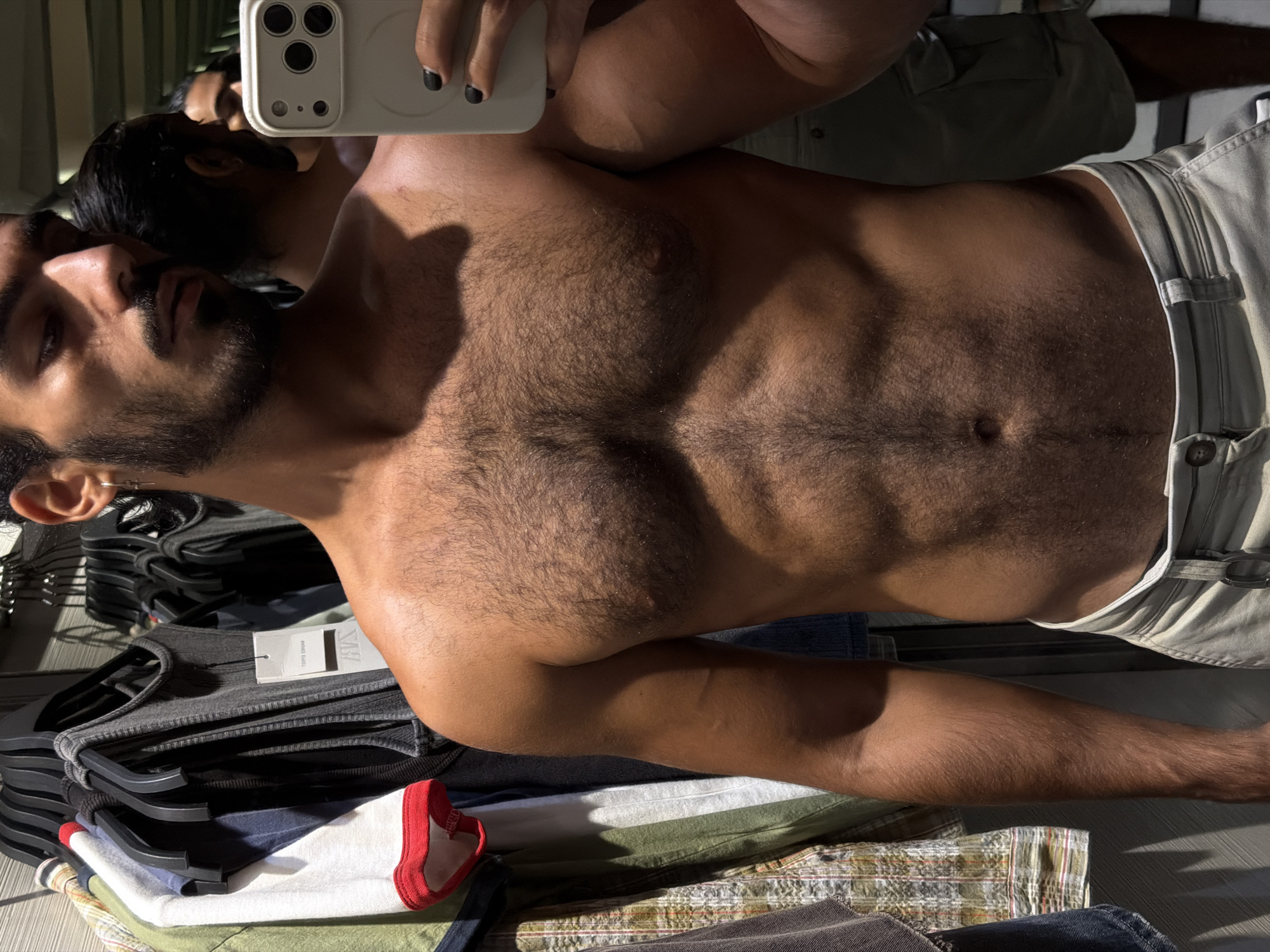 A man with dark hair and a beard takes a mirror selfie in what appears to be a clothing store. He is shirtless, revealing a hairy chest and abs, and is wearing light-colored shorts. In the background, racks of clothes are visible. The reflection in the mirror shows another man, also shirtless. The lighting suggests it might be daytime. This scene is located in the city of Kuta, Indonesia.