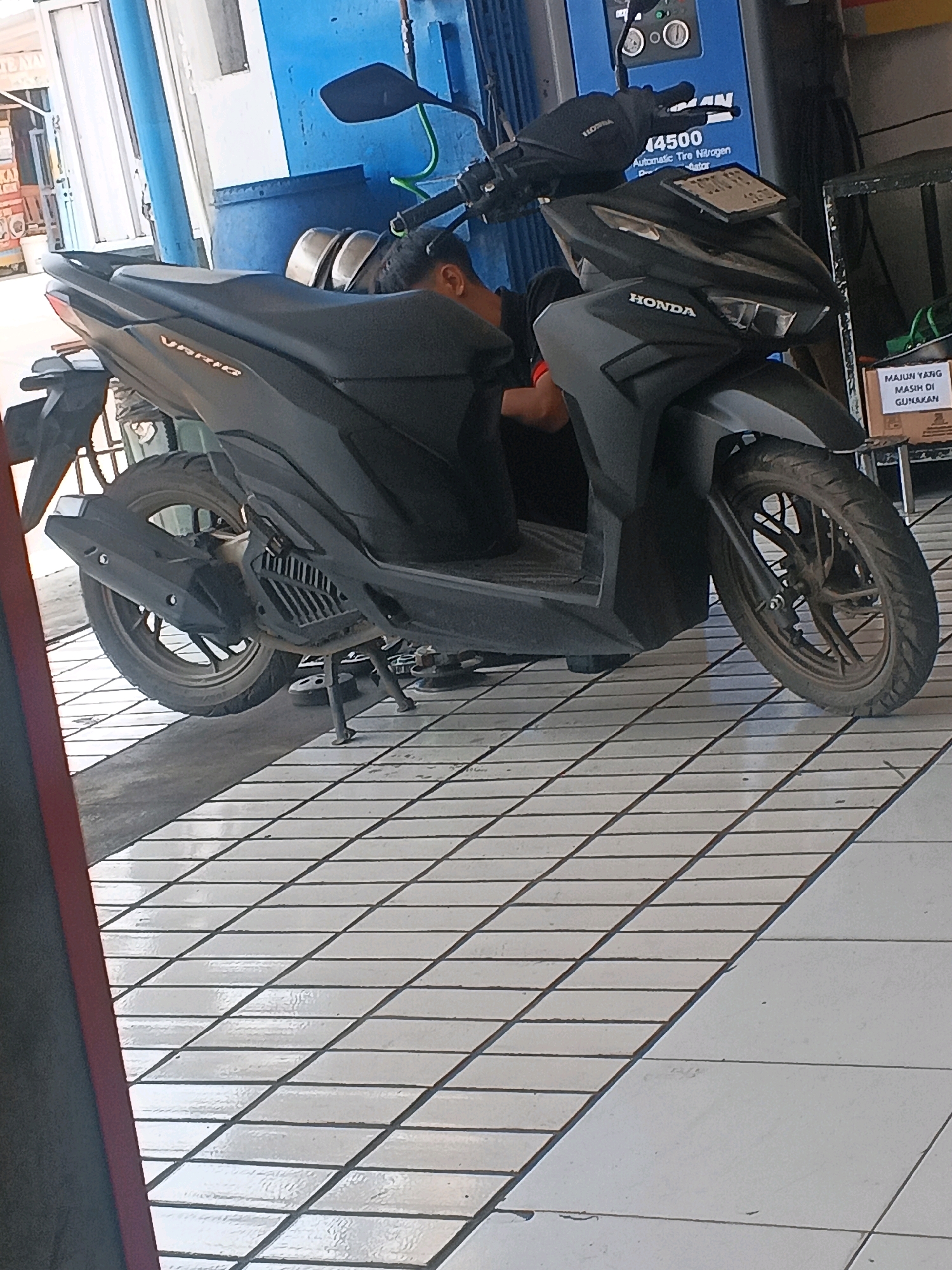 In Taman, Indonesia, a person is seen in a kneeling position next to a dark gray Honda Vario scooter. The scooter is parked on a tiled floor, with diagonal white tiles forming a grid pattern. Behind the scooter, a blue machine with gauges and text is visible, possibly a tire inflation station. A small cardboard box with Indonesian text, "MAJU YANG MASIH DI GUNAKAN," is placed to the right of the scooter. The lighting suggests it is daytime, with sunlight casting shadows on the floor.