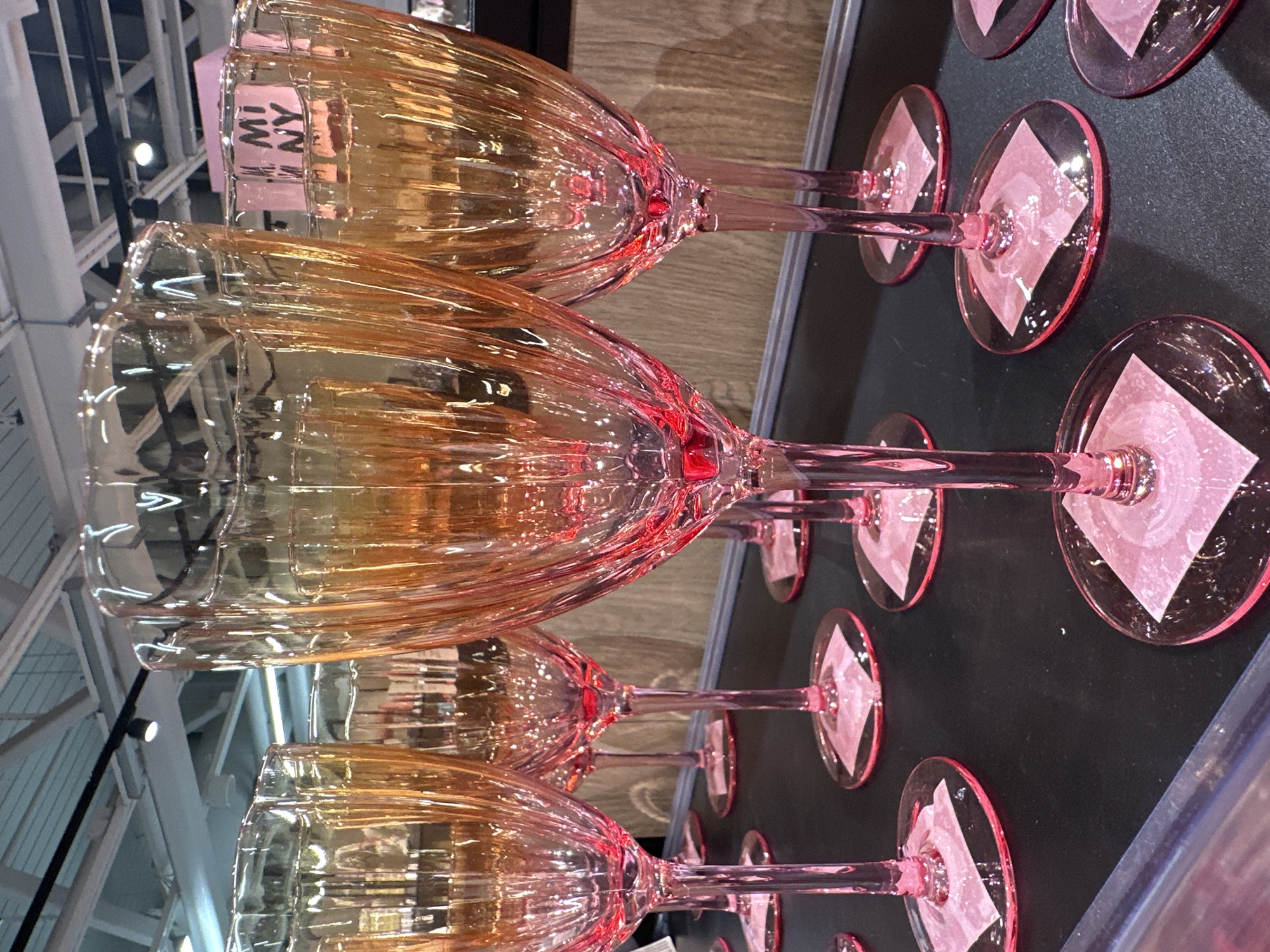 This media file displays a close-up, angled view of several tinted glassware items, likely wine glasses, arranged on a dark surface. The glasses are a gradient of amber and pink hues, with a ribbed texture on the bowl. The stems and bases of the glasses are a translucent pink. Each glass has a small, light pink paper insert, possibly a price tag or protective material, placed on its base.
In the upper left corner of the image, a label with black lettering on a pink background is visible within one of the glasses. The visible text reads "MI NY". The background behind the glasses consists of a wooden texture and elements of a retail environment, including what appear to be ceiling structures and lighting. The scene is well-lit, suggesting an indoor setting like a store or display area. No people are visible in the frame, and the activity taking place is simply the display of these decorative items. The overall impression is one of a product showcase.