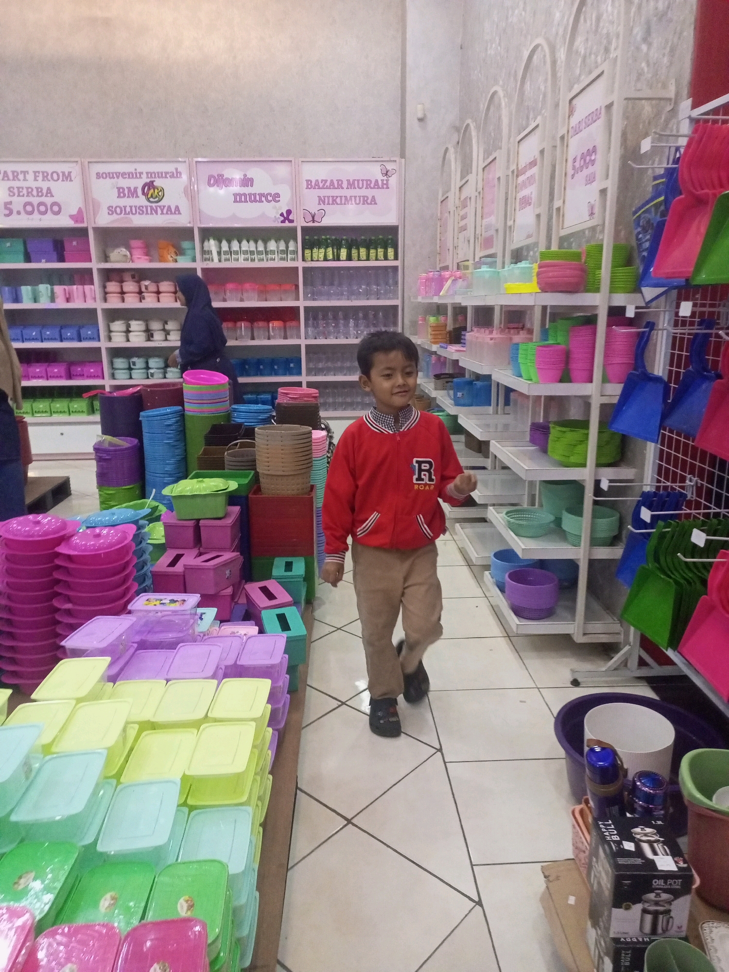 The image depicts the interior of a retail store, likely a homeware or general goods shop, situated in Ngasem, Indonesia. In the foreground, a young boy, appearing to be around 6-8 years old, walks towards the right of the frame. He is wearing a red jacket with white trim and an emblem on the left chest, paired with tan pants and dark shoes. He is looking towards the camera with a neutral expression.
The store is filled with merchandise, predominantly plastic containers and kitchenware, displayed on white shelves and in stacks on the floor. The shelves are organized with rows of colorful plastic bowls, cups, and storage boxes in various shades of pink, purple, green, yellow, and blue. In the background, a woman wearing a dark blue hijab is visible, interacting with the merchandise on a shelf.
Promotional signs are mounted on the wall above some of the shelves. One sign reads "ART FROM SERBA 5.000," another "souvenir murah BM," and another "Dijamin murce." Further down the aisle, more shelves are packed with similar items. On the right side of the image, the aisle widens, showing a display of colorful plastic dustpans and brooms. On the floor in the lower right corner, several larger items are visible, including buckets and boxed products, one of which is labeled "OIL POT." The flooring is a tiled pattern in white and light gray. The lighting appears to be artificial, illuminating the store environment.