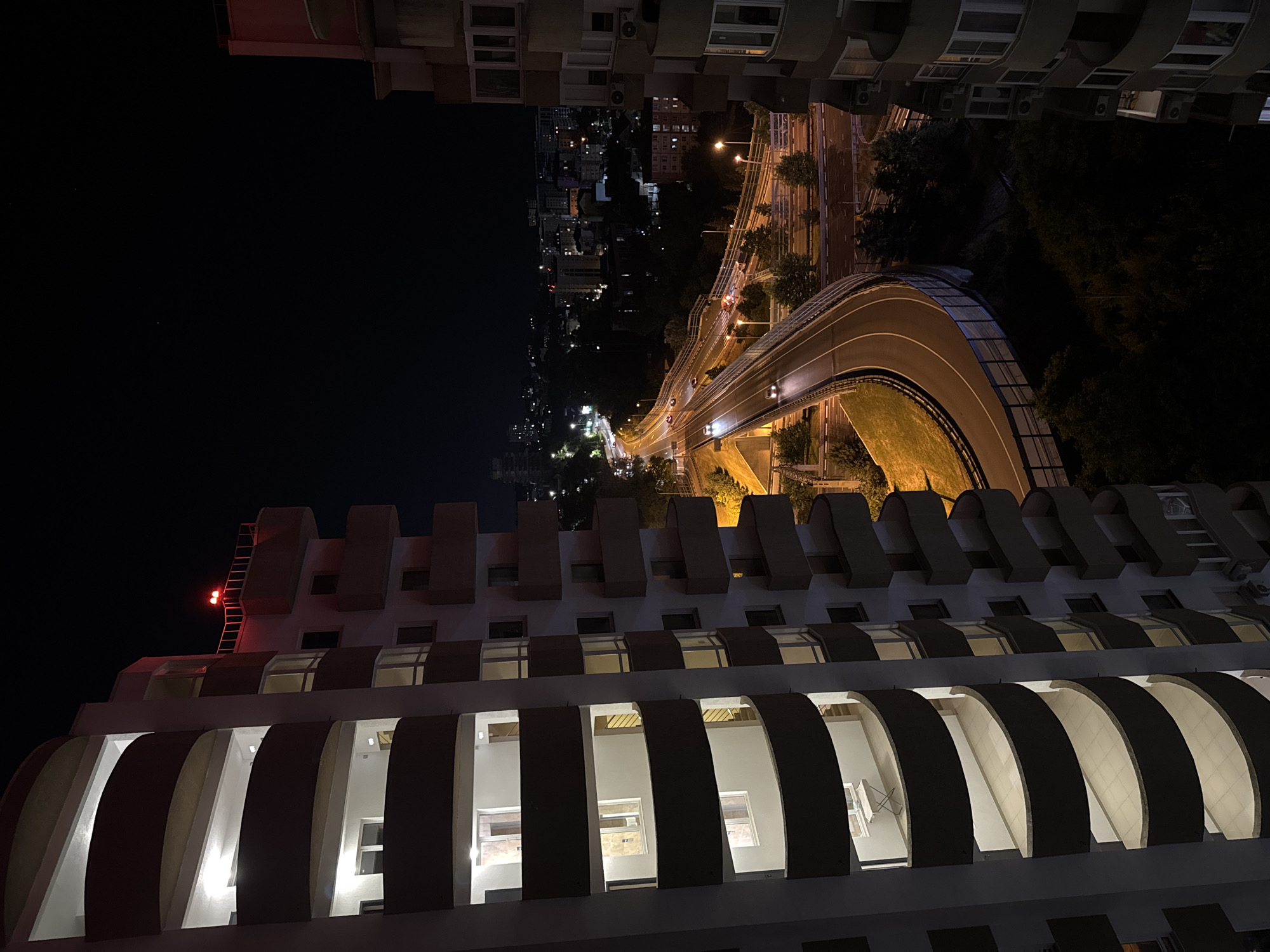 The image depicts an aerial view of a city at night, focusing on a multi-lane road curving through an urban landscape. The scene is dominated by the artificial lights of buildings, streetlights, and vehicles.
On the left side of the frame, a tall residential building with curved balconies is prominently featured. The balconies are illuminated from within, revealing some empty spaces and the distinct architectural style of the building. On the upper part of this building, a red warning light is visible on a railing.
To the right of the road, another building with a more angular design and smaller, rectangular balconies is partially visible. These balconies also show interior lighting.
The road itself is illuminated by streetlights, highlighting the asphalt surface and traffic lanes. Several cars are visible on the road, their headlights and taillights creating streaks of light. The road appears to be elevated or part of a complex interchange, with a metal railing and a sound barrier separating it from the surrounding area.
Beyond the immediate road and buildings, the cityscape extends into the distance, with numerous buildings of varying heights and illumination levels. The overall impression is one of a bustling city at night, with a focus on transportation infrastructure and residential architecture.
The image was taken in the city of Sochi, Russia, likely from a high vantage point, possibly another building or a drone. The time of day is night, indicated by the dark sky and the reliance on artificial lighting. No people are explicitly visible or performing any actions. The environment is urban, characterized by concrete structures, asphalt roads, and some areas of greenery, which are largely obscured by darkness.
