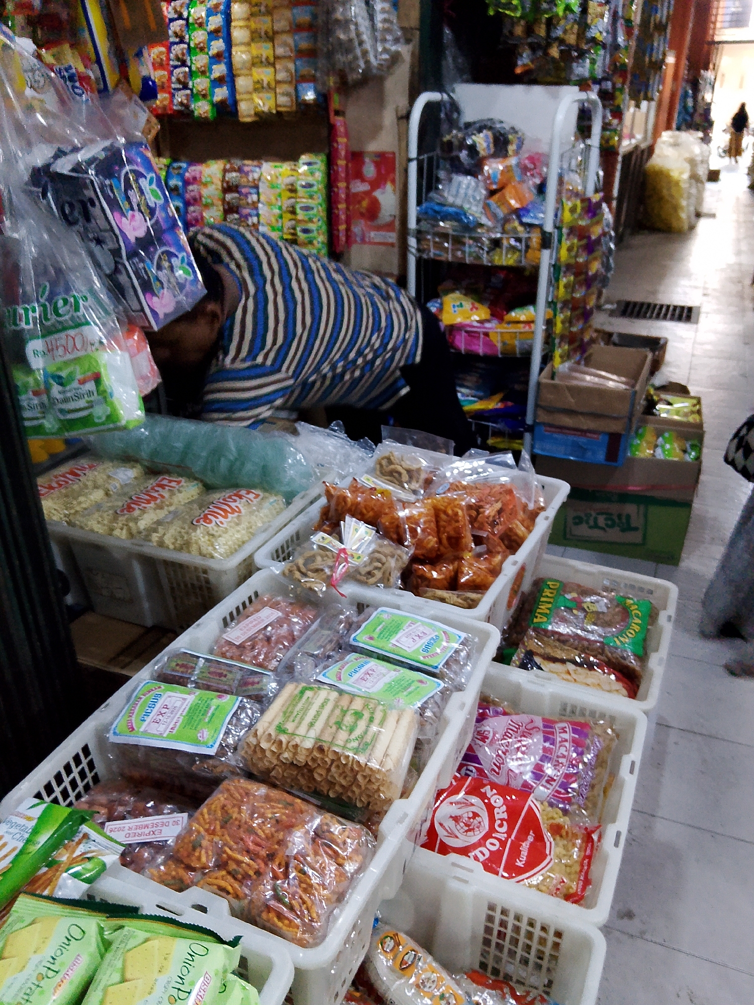 The image depicts a street market stall in Tanubayan, Indonesia, filled with various packaged snacks and food items. A person, partially visible, is seen bending over a display of goods, appearing to be organizing or attending to the products. The stall is densely packed with colorful packaging, including bags of chips, crackers, and other assorted snacks, neatly arranged in white plastic baskets. Behind the stall, shelves are stocked with more merchandise, and in the background, the market aisle is visible, with other stalls and shoppers in the distance. The lighting suggests it is daytime, and the overall atmosphere is that of a busy, local marketplace.