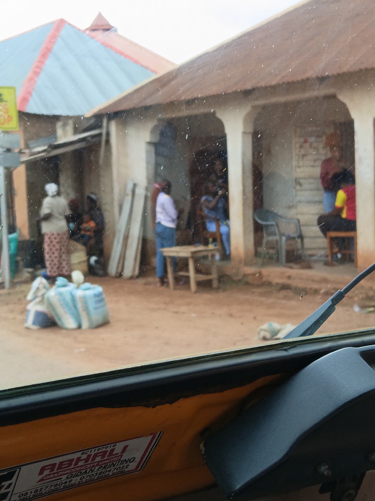 The image shows a street scene in front of a building with a corrugated metal roof. Several people are visible, some standing and some sitting. In the foreground, there are several large sacks or bundles on the ground, as well as a part of a vehicle, likely a tricycle or auto-rickshaw, with a yellow interior visible. The building appears to be a shop or a dwelling, with an open front revealing the interior. The ground is dirt or sand. There are also some chairs visible outside the building. The overall impression is of a bustling, everyday scene in a warm climate.