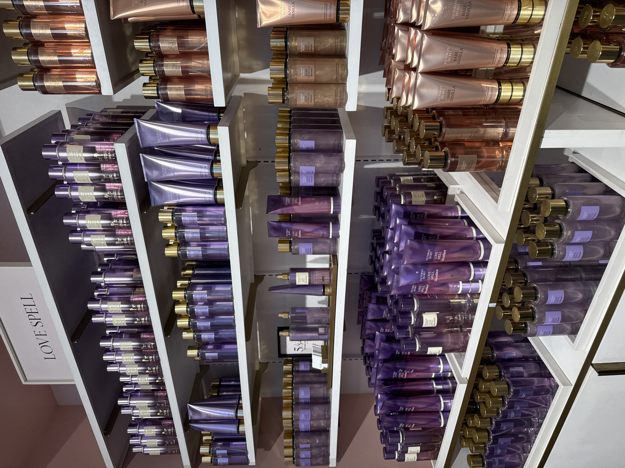 The image shows a retail display of Victoria's Secret body care products, primarily lotions and body mists, in varying shades of purple and rose gold. The products are arranged on multiple white shelves against a pink wall. A white sign with black text reading "LOVE SPELL" is prominently displayed at the top of the shelving unit. The products appear to be organized by type and scent, with numerous bottles and tubes neatly lined up. The overall impression is one of a well-stocked and organized retail environment. The specific location of this store in New York City is not discernible from the image itself.