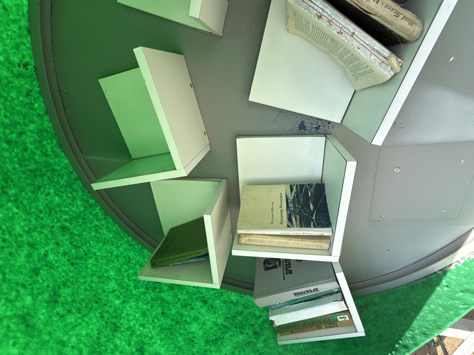 A top-down shot of a circular public bookcase in the city of Синьково, Russia, filled with Russian-language books of various sizes. The bookcase is painted gray with white shelves. Each shelf contains at least one book, with some containing stacks. The ground around the bookcase is covered in bright green artificial turf. One book in the bookcase has a damaged spine. Handwritten writing is faintly visible on the side of the bookcase.