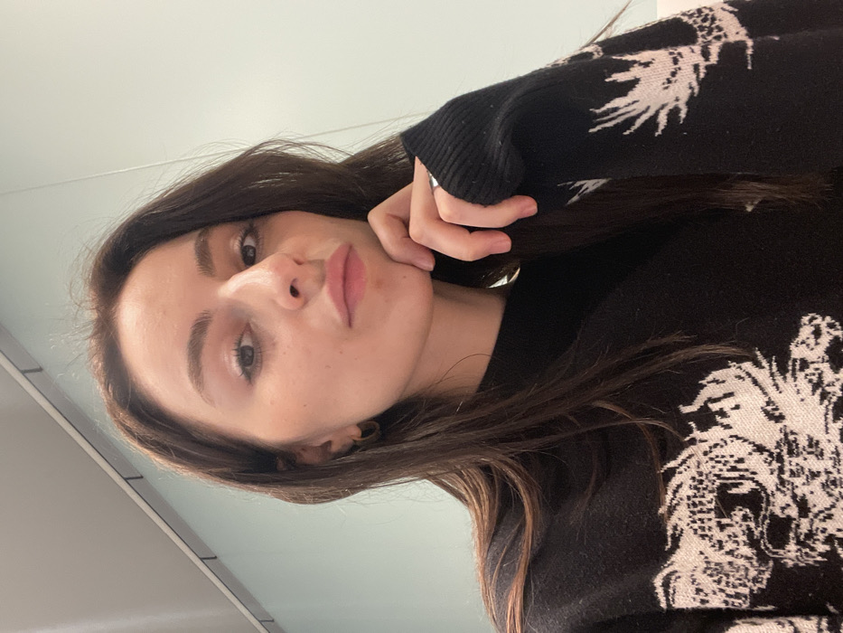 A young woman with long, dark brown hair poses in a selfie. She wears a black sweater with a white design on the sleeves. Her hand rests on her chin. She looks up at the camera with light eyes. Her lips are pink and full. The background is plain and light-colored. The image is slightly tilted.