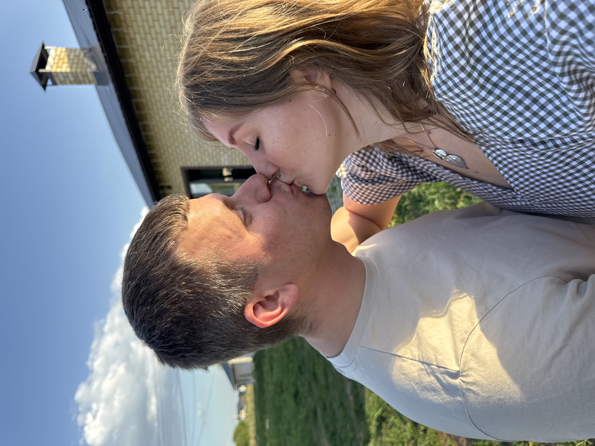 A couple is kissing outdoors in Ферма, Russia. The woman has light brown hair and is wearing a checkered dress. The man has short dark hair and is wearing a light-colored t-shirt. Behind them is a building with a chimney and a lawn with green grass. The sky is visible in the background with some clouds.