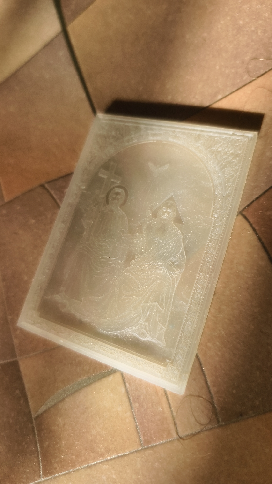 This image shows a close-up, angled view of a rectangular, translucent object with a raised relief image on its surface. The object is resting on a tiled floor with alternating shades of brown and beige square tiles. The lighting casts a strong shadow from the object onto the floor.
The relief image depicts two figures seated side-by-side within an arched frame. The figure on the left appears to be standing and is partially obscured by the seated figure. This figure holds a cross aloft, and has a halo around its head. The figure on the right is seated and appears to be a more central figure. A dove is depicted in the upper center of the arch, above the figures. The relief is detailed, showing drapery on the figures' clothing and decorative patterns around the edge of the arched frame. The material of the object is slightly opaque and has a light, creamy color.
The scene appears to be religious or devotional in nature, possibly an icon or a plaque. The lighting suggests it is daytime, but there are no specific indicators of weather or the exact time of day. The setting is indoors, on a floor with prominent grout lines between the tiles. There is a single strand of dark hair visible on the tiled surface to the right of the object. No text is visible in the image.
