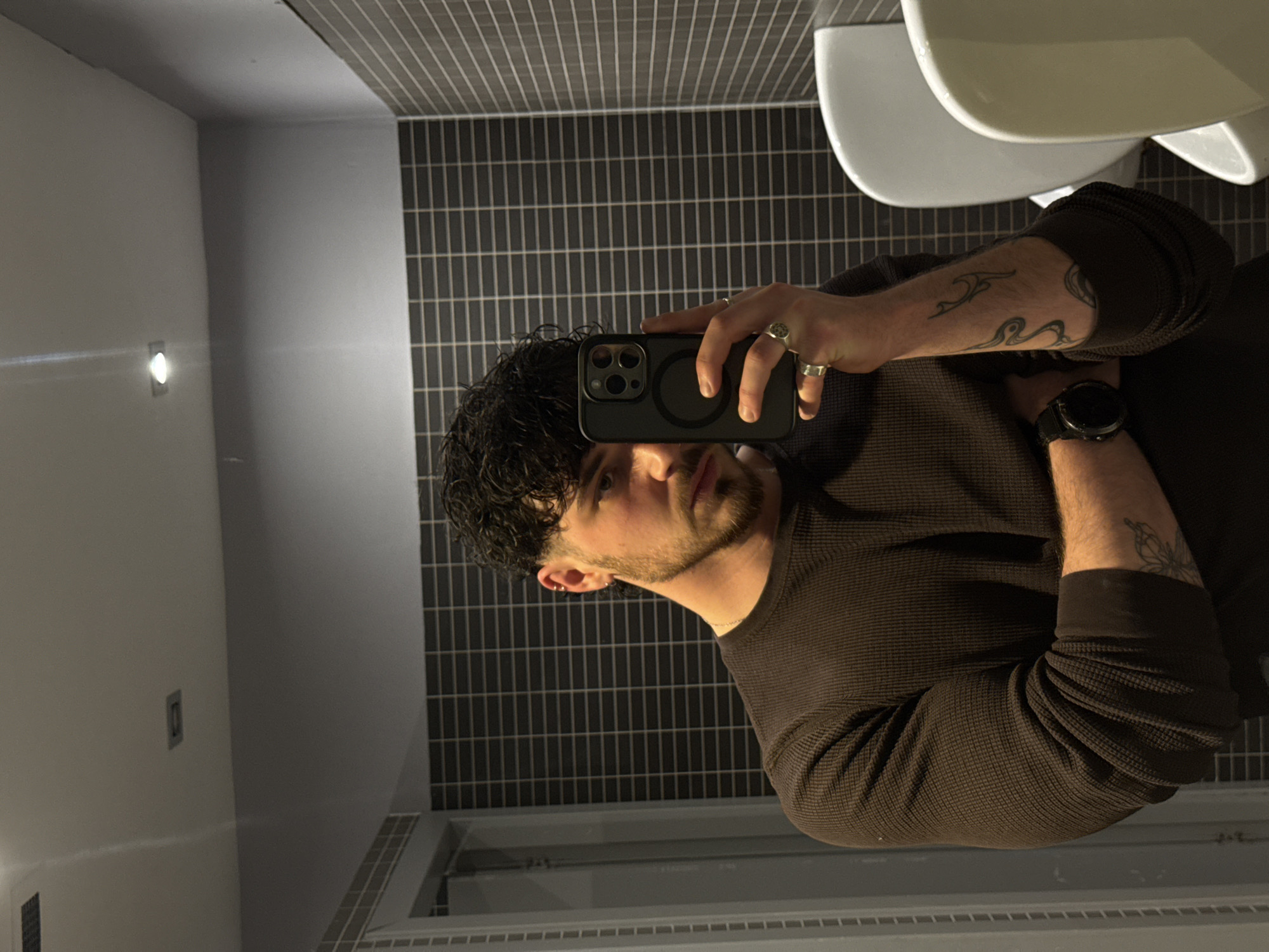 A man is taking a mirror selfie in a bathroom. The wall behind him is covered in dark gray, horizontally laid, rectangular tiles. A white urinal is visible in the upper right corner of the frame. The man has dark curly hair, a beard, and is wearing a long-sleeved brown waffle-knit shirt. He is wearing a watch on his left wrist and several rings on his fingers. The phone he is using has a black case. The bathroom has a white door frame and molding along the bottom of the wall.