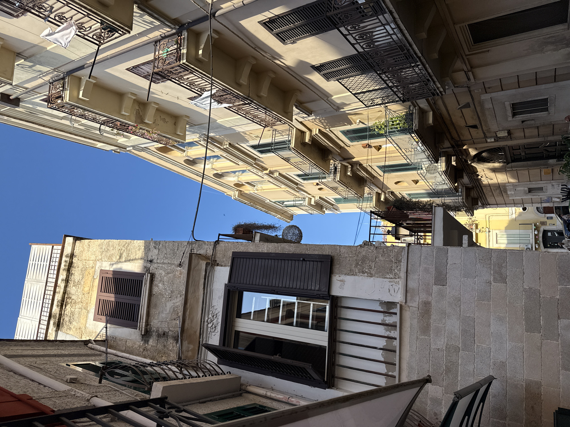 The image presents a worm's-eye view looking upwards into a narrow urban space, framed by densely packed multi-story buildings under a clear, bright blue sky. The scene is illuminated by strong sunlight, creating distinct areas of light and shadow across the facades.
The buildings display a mix of architectural styles. On the left, several structures are painted in lighter, warmer tones, possibly pale yellow or cream. These feature ornate wrought-iron balconies, some adorned with potted plants, and one with white laundry or linen hanging to dry. Windows with dark shutters, some closed and some open, are visible on these buildings.
On the right, the buildings appear older, constructed from weathered gray stone or concrete blocks. These also feature windows with dark shutters, some half-open, reflecting parts of the opposite buildings or the sky. Intricate iron railings and grates are present on balconies and windows. Various utility lines and cables crisscross the space between the structures. Notable details include a decorative floral carving on a wall and a spherical, woven metal art piece placed on a high ledge.
No people are visible in the image, but the presence of hanging laundry and open windows suggests active habitation. The clear sky and strong shadows indicate it is daytime, likely with the sun high in the sky. The architecture and density evoke the feeling of an old city, possibly in a Mediterranean or European region. There is no visible text in the image.