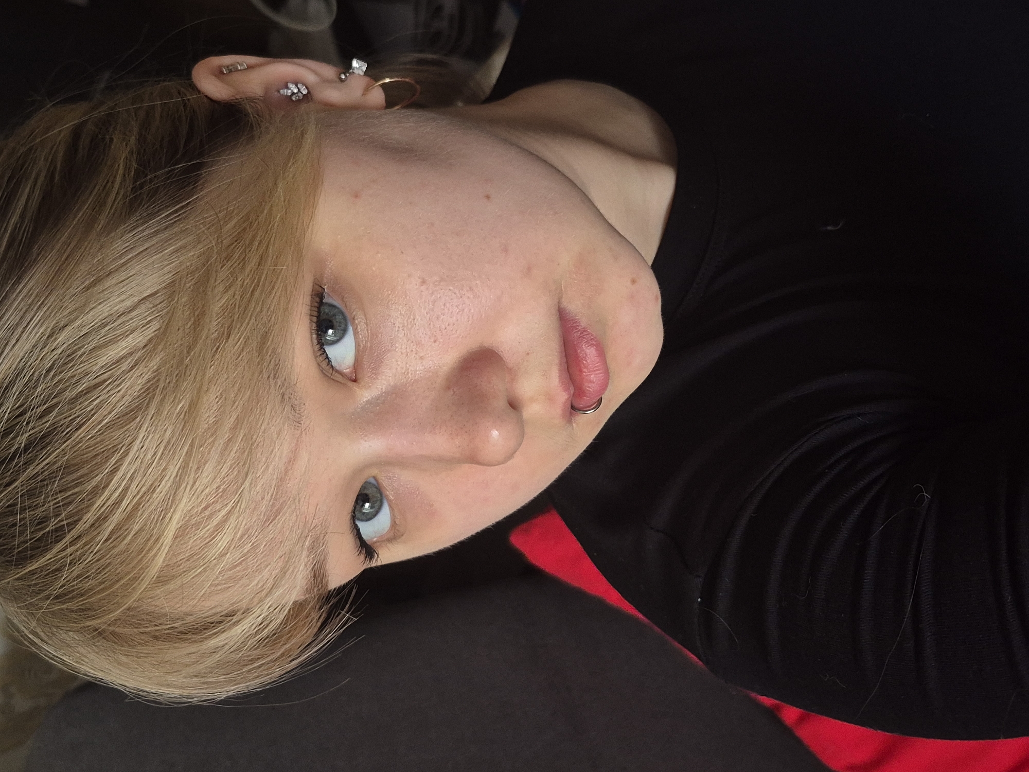 A young Caucasian woman with light blonde hair and blue eyes looks upward at the camera. She has multiple piercings in her left ear and a lip ring. Her hair is styled with a side-swept fringe. She is wearing a black top with a hint of red visible near the neckline. The background is dark and indistinct. It is not possible to identify the location in the city of Chaikovsky, Russia from the provided image.