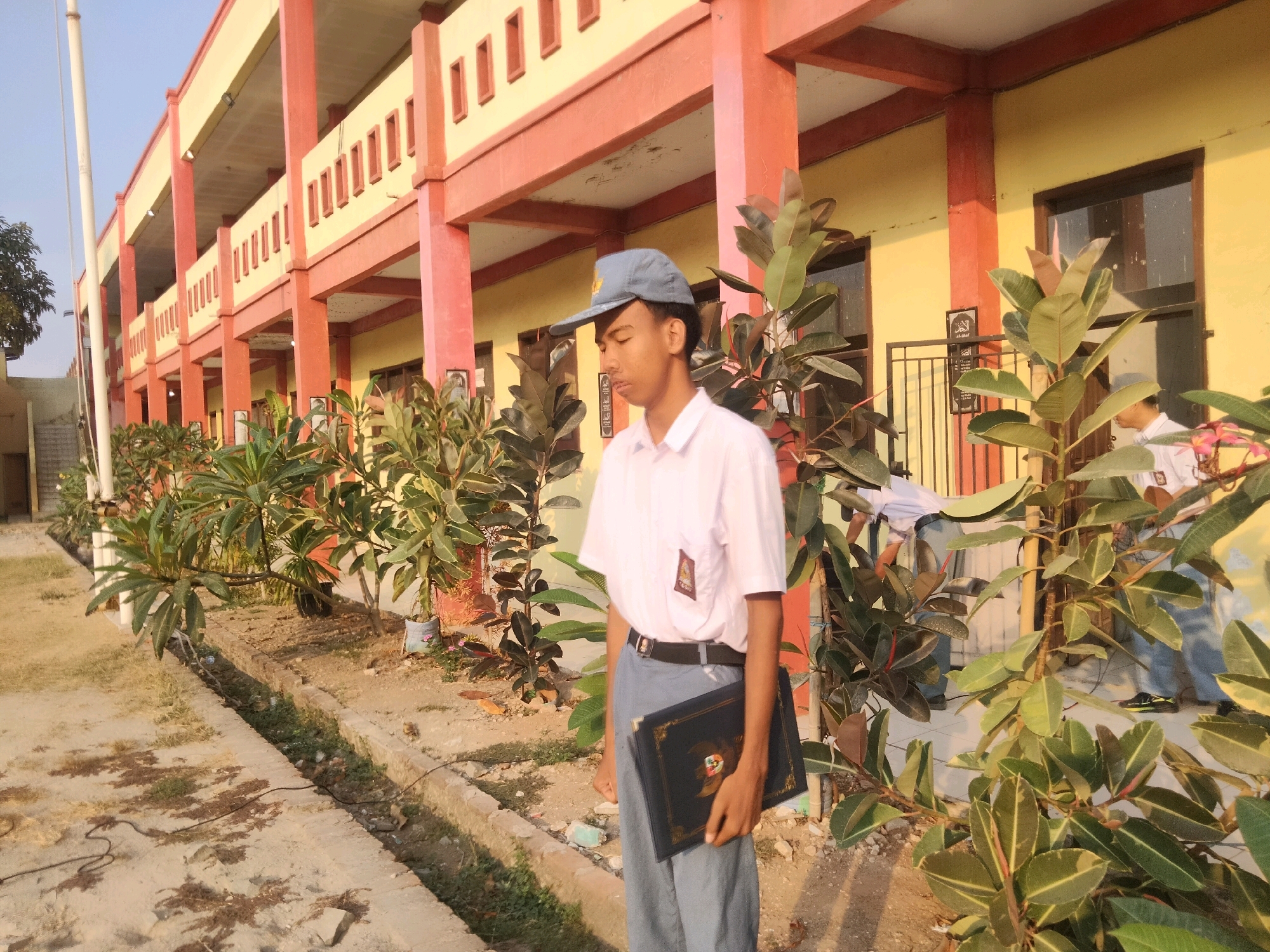 In the city of Suka Sari, Indonesia, a young person wearing a school uniform, including a white collared shirt, blue pants, and a blue cap, stands in front of a two-story building. The building has a yellowish facade with orange trim around the windows and pillars supporting an upper-level balcony. The young person is holding a dark-colored book with gold embellishments. In the background, other individuals in similar school uniforms are visible near the building. The area in front of the building is lined with several potted plants and trees with large, glossy leaves. The ground is a mix of dirt, sand, and some patchy grass, with a concrete path running alongside the plants. A drainage ditch is present near the path. The lighting suggests it is late afternoon, with the sun casting warm shadows.