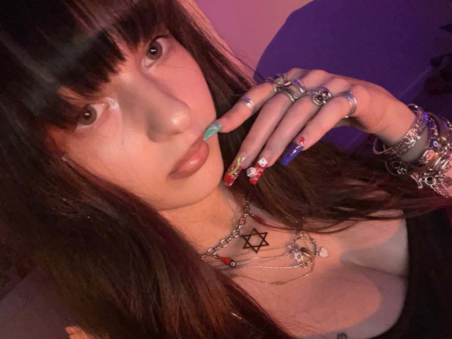A woman poses closely for the camera, displaying a variety of accessories. She wears multiple rings on her fingers and several bracelets on her wrist. Her nails are long and elaborately decorated. Around her neck, she wears layered necklaces with a Star of David. She has dark hair with bangs, fair skin, and light brown eyes. The lighting appears to be a combination of pink and purple hues.