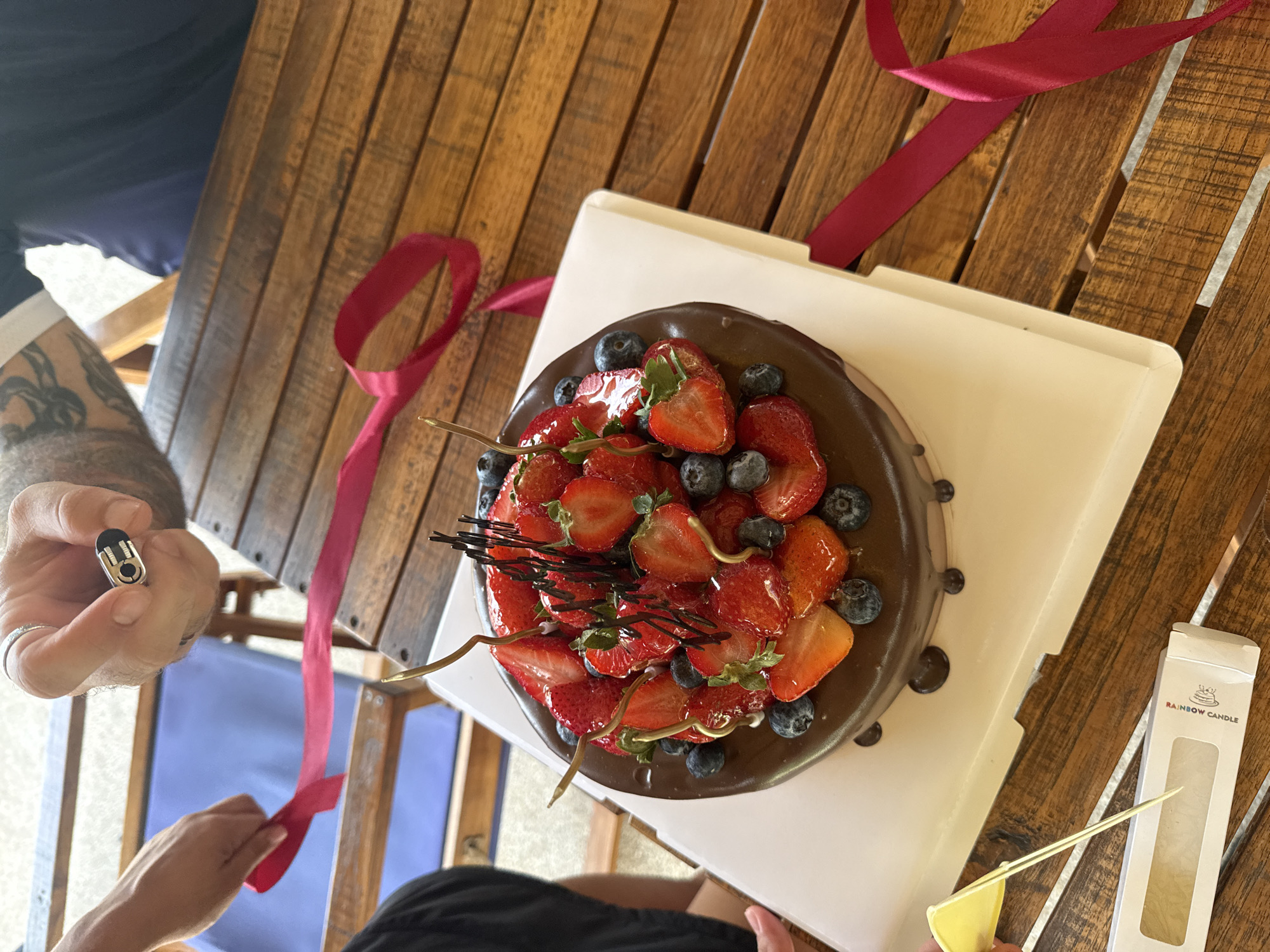 A high-angle shot shows a chocolate cake topped with strawberries and blueberries on a white cake board. The cake is on a wooden table. To the left, a hand holds a red ribbon. In the upper right, a person holds a small silver object near their hand, which wears a ring. The environment suggests an outdoor setting, possibly a cafe or restaurant patio, with a glimpse of blue seating in the background. The table is made of dark wood planks. A small box with "RAINBOW CANDLE" printed on it is in the lower left. The location is identified as Baan Bang Rak, Thailand.