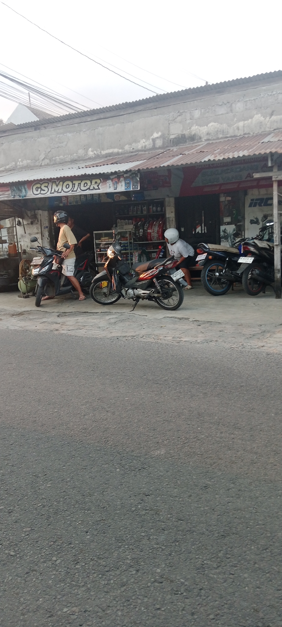 A side view of the exterior of a motorcycle repair shop and storefront in Maguwoharjo, Indonesia, displays a scene with several motorcycles and people. The shop, identified by signage reading "GS MOTOR," has a corrugated metal roof and a concrete facade. Shelves stocked with various items, including oil bottles and tires, are visible inside.
In the foreground, a person wearing a light brown t-shirt and shorts, with a black helmet, stands next to a dark-colored motorcycle. To their right, another dark-colored motorcycle is parked. Further to the right, a person wearing a white shirt and a white helmet is bent over a red and black motorcycle. Behind this motorcycle, a darker colored motorcycle with a blue rim is parked. The ground in front of the shop is a cracked concrete surface, transitioning to asphalt at the bottom of the frame. The sky is overcast with visible power lines stretching across the top of the image.
