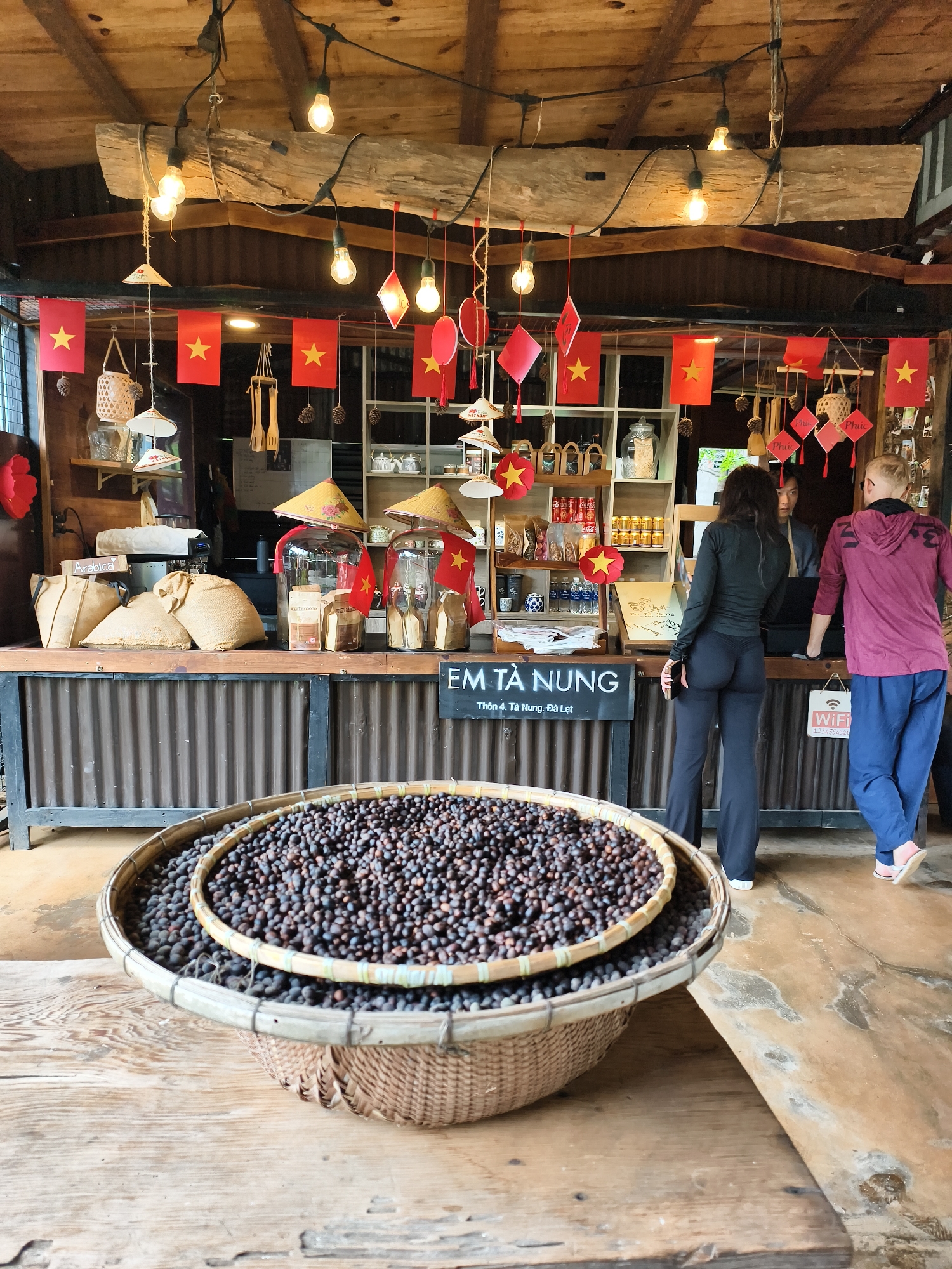 The image captures a scene inside what appears to be a coffee shop or store in Thôn 4, Tà Nung, Đà Lạt, Vietnam. The interior has a rustic aesthetic, with a wooden roof structure adorned with exposed light bulbs and Vietnamese flags. The shop counter is made of corrugated metal. Behind the counter, shelves display various items including coffee beans, jars, and other merchandise. Two customers are at the counter, one woman wearing a black top and pants, and a man in a maroon hoodie and blue pants. In the foreground, a large woven basket is filled with what looks like coffee beans. A sign on the counter reads "EM TÀ NUNG" and mentions the location as Thôn 4, Tà Nung, Đà Lạt, as well as a Wi-Fi password.