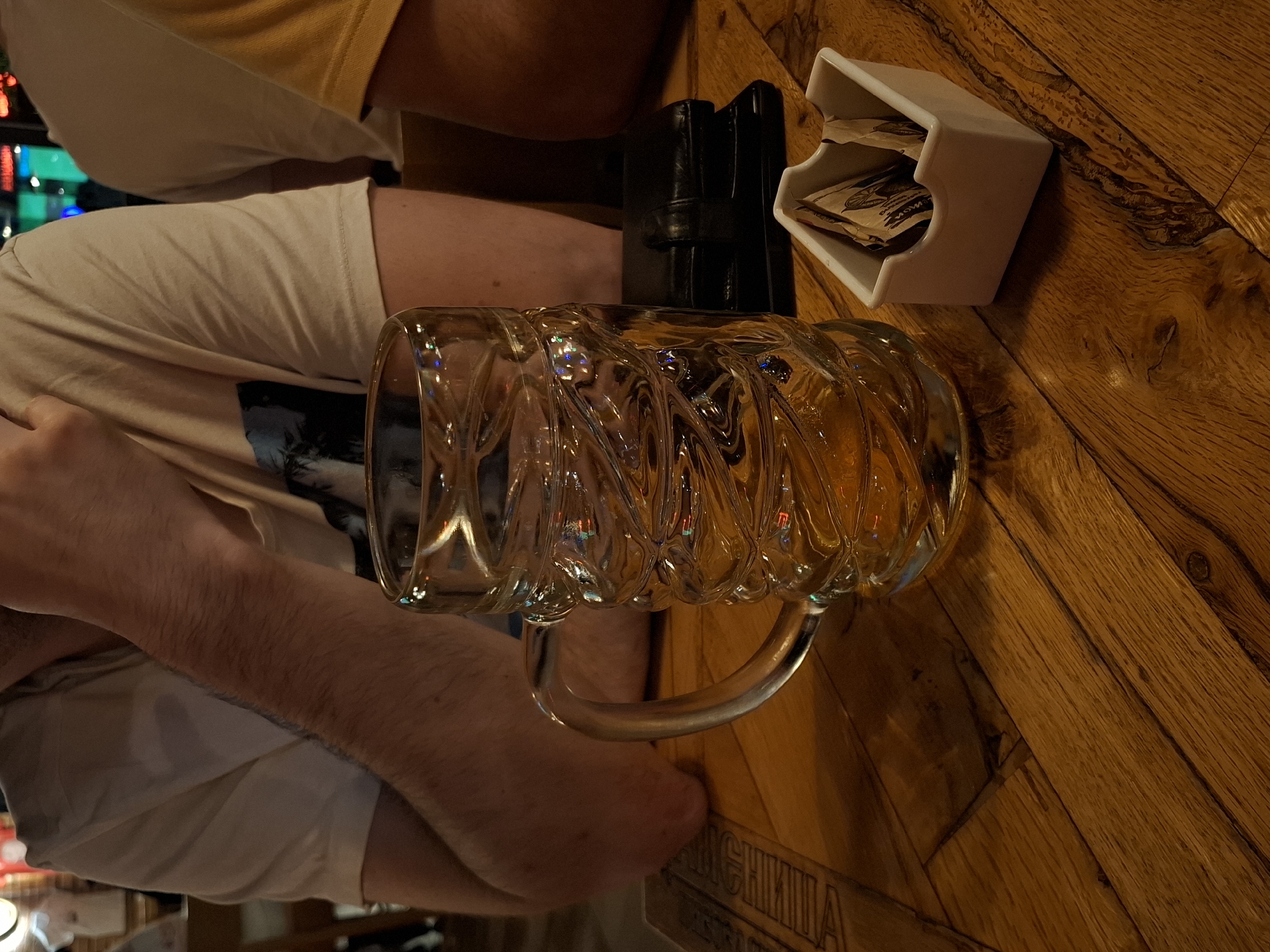 A glass mug filled with a golden liquid sits on a wooden table. The mug has a textured glass design and a sturdy handle. To the right of the mug, a small white dish contains several packets of sugar. Behind the mug, a black wallet is placed on the table. The table surface is made of wooden planks. In the background, parts of two individuals are visible, wearing light-colored t-shirts. The overall setting appears to be an indoor establishment, likely a bar or restaurant, with a warm, ambient lighting.