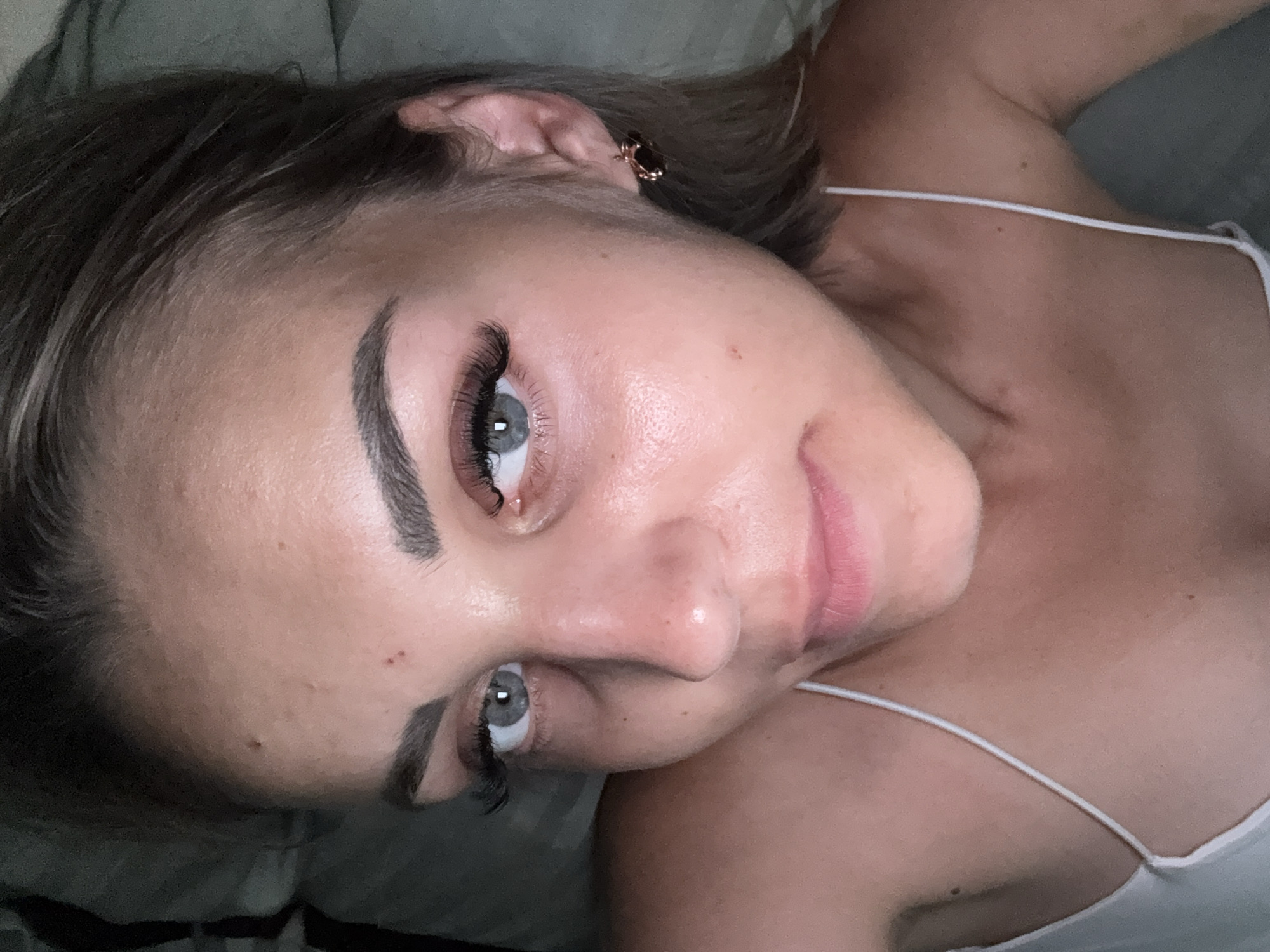 A close-up, top-down selfie of a young woman with light skin, fair hair, and light blue eyes is captured in a dimly lit indoor setting. She is wearing a white camisole with thin straps. Her face is the primary focus, with dramatic false eyelashes enhancing her eyes. Her eyebrows are well-defined, and she is wearing subtle makeup. A small mole is visible on her forehead, and there are a few other marks on her skin. She has a small earring in her right ear, appearing to be a dark stone set in metal. The background is dark and out of focus, suggesting a bedroom or similar private space. The image does not contain any elements that would suggest a specific location in Yuzhno-Sakhalinsk, Russia, or any other city.