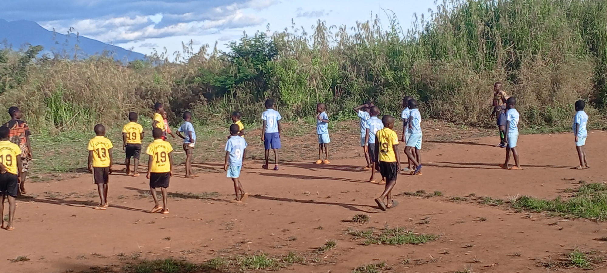 The image depicts a group of children in Ngalula, Democratic Republic of the Congo, on a dirt field. Most are wearing sports jerseys, some yellow with "19" on the back, and others light blue with "9". The children are dispersed across the field, with some appearing to be in conversation or standing around. The background includes tall grasses and bushes, as well as a mountain range visible in the distance under a cloudy sky. The overall environment suggests a rural or semi-rural setting.