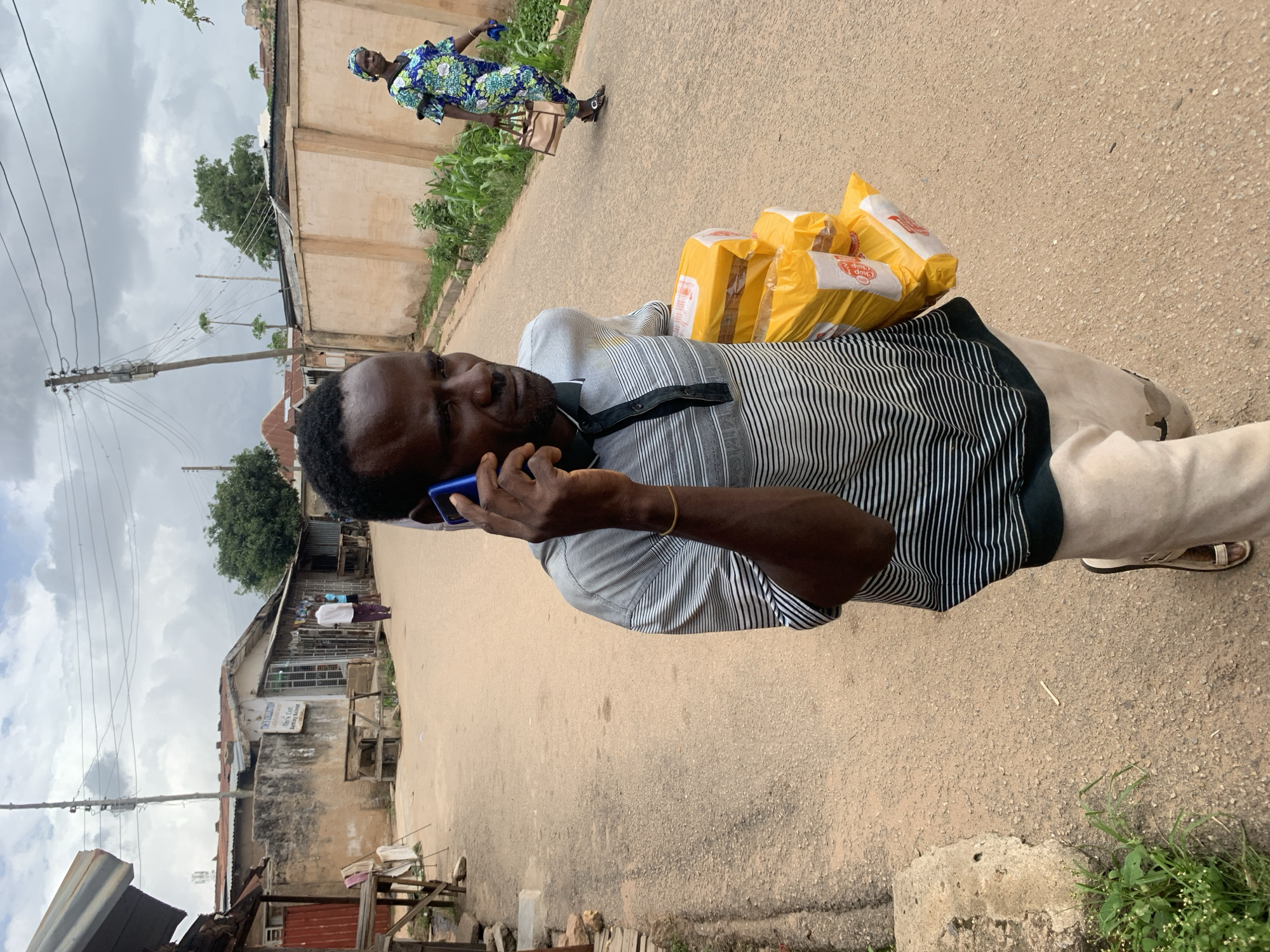 A man stands on a paved street in Jos, Nigeria, holding a blue cell phone to his ear with his left hand. He carries multiple yellow bags over his left shoulder. He wears a striped shirt with black and white vertical stripes and khaki pants. In the background, a woman in a patterned blue dress walks along a paved sidewalk next to a tall, beige-colored wall. A building with a corrugated metal roof stands to the left, with power lines visible above. The sky is cloudy.