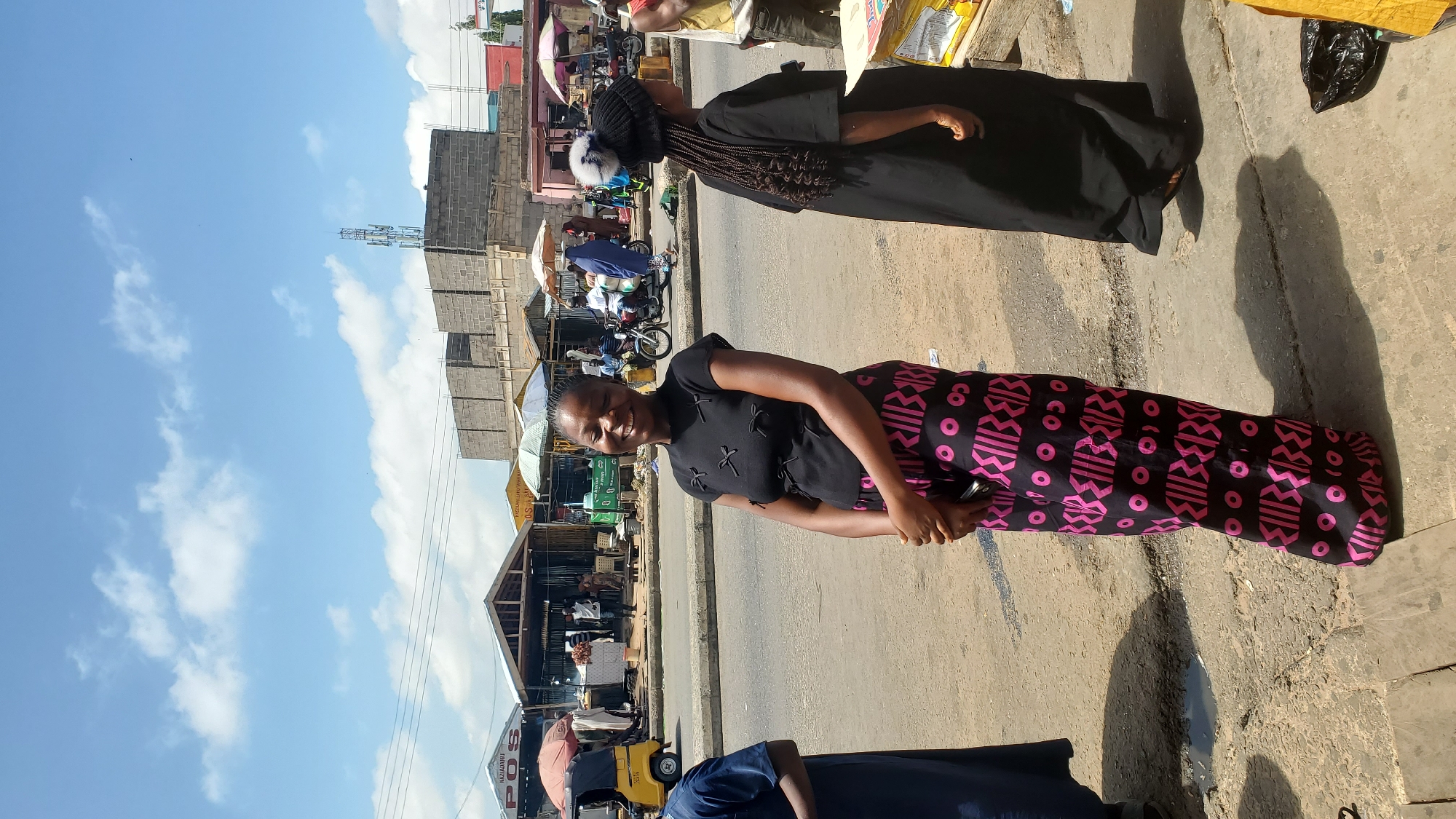 This is a candid, eye-level shot of two women in what appears to be a bustling outdoor market. The image is oriented vertically. The woman in the foreground is smiling widely and looking directly at the camera. She is wearing a black short-sleeved top adorned with small black bow details and a long, flowing skirt with a vibrant pink and black geometric pattern. She is holding a dark-colored smartphone in her right hand. Behind her, another woman is partially visible, wearing a black garment and a black head covering with a white pom-pom. Her hair is styled in braids that hang down her back.
The setting is an outdoor market with several stalls and structures visible in the background. There are various people and motorcycles scattered throughout the scene, suggesting a lively atmosphere. Sunlight casts distinct shadows on the ground, indicating it is daytime, likely midday due to the angle of the shadows. The sky is bright blue with scattered white clouds, suggesting fair weather. One of the stalls has signs with illegible text.