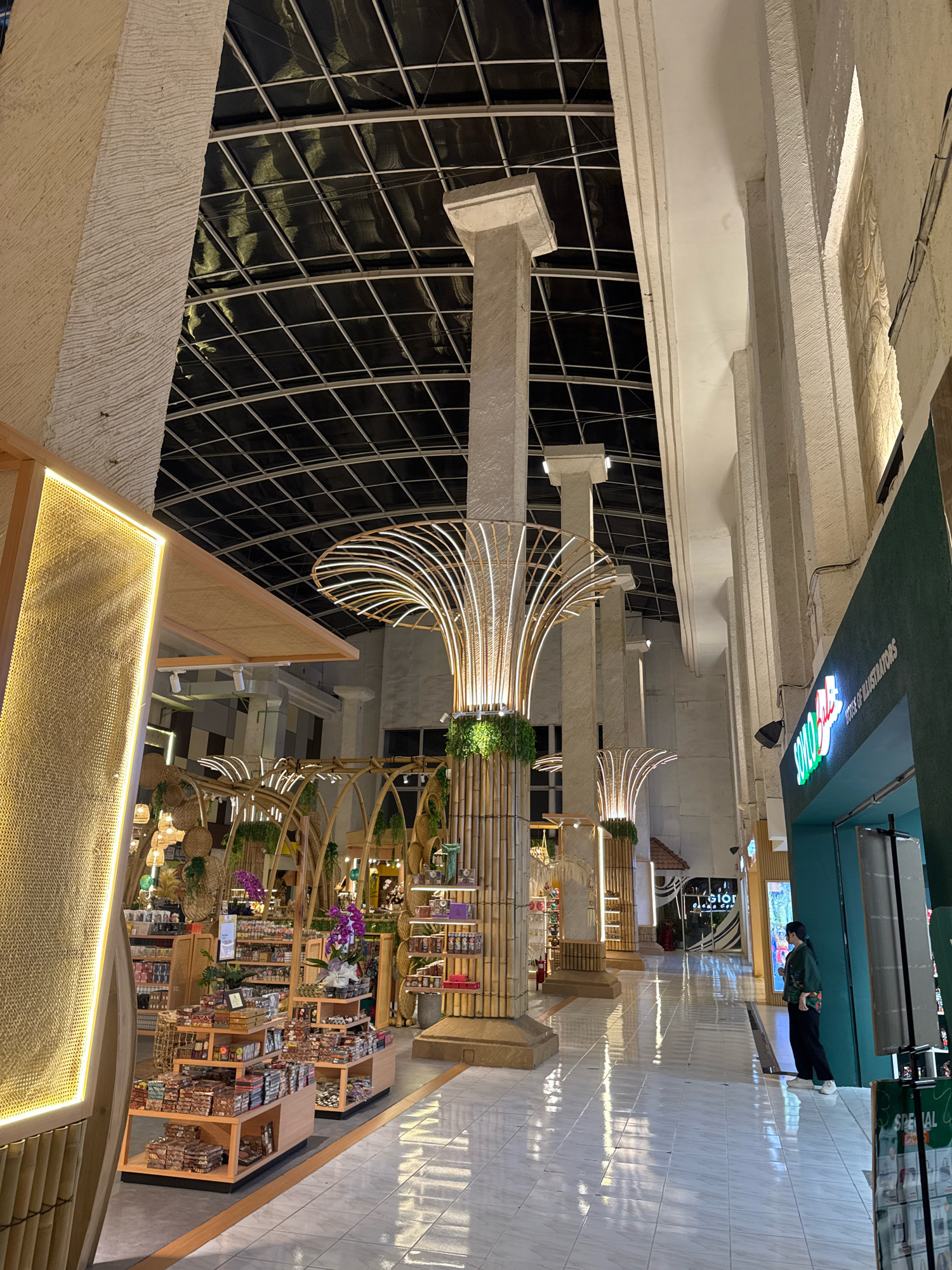 The image depicts the interior of a marketplace in Kuta, Indonesia. The space features a high, arched ceiling with a black fabric covering supported by a metal grid structure. Large, textured concrete pillars line the walkway, some adorned with decorative bamboo elements and lush green plants. The walkway itself is paved with white tiles that reflect the ambient lighting.
On the left side of the image, a section of a store is visible, showcasing merchandise on wooden shelves. The store facade is decorated with a woven, textured material illuminated by a warm yellow LED light strip. Further into the marketplace, more stalls are visible, displaying an assortment of products on tiered wooden stands. These products appear to be packaged goods, possibly food items, with some also featuring decorative orchids.
In the background, a store sign reads "SOVO HOUSE OF ILLUSTRATORS" in green lettering on a teal background. A woman, dressed in a dark green jacket and black pants, stands facing the store, looking at a display. The overall ambiance is warm and inviting, with the lighting creating a sense of depth and highlighting the natural materials used in the decor.
