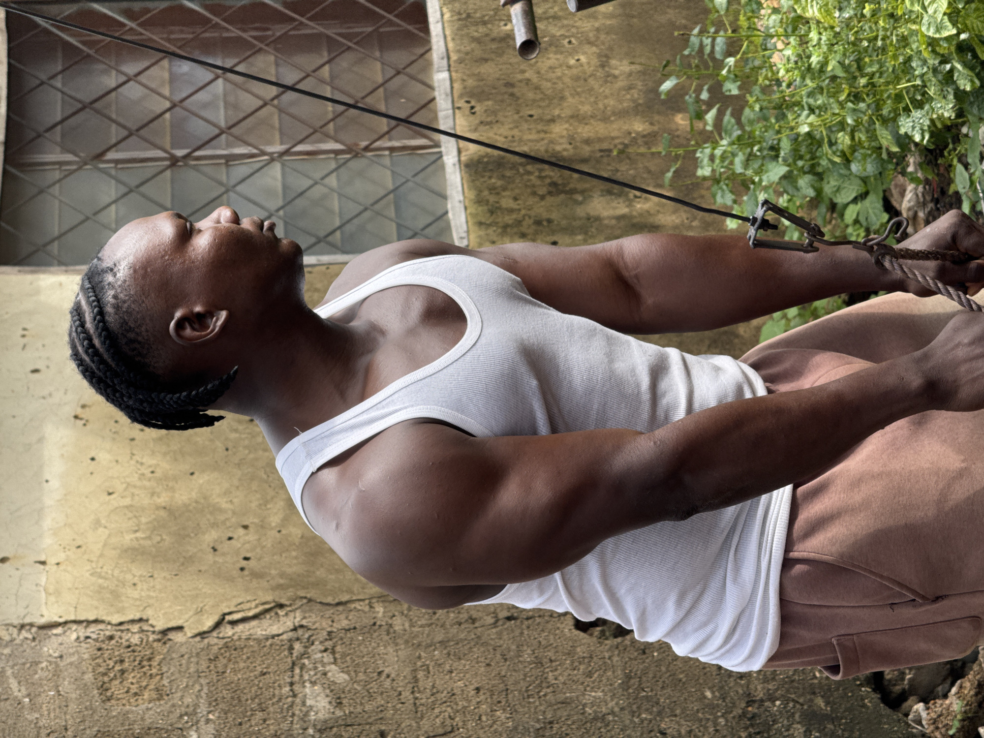 A person is captured from the chest up, in a close-up shot taken outdoors. The individual, likely a man given the visible muscular physique and braided hair, is wearing a white ribbed tank top and brown sweatpants. Their head is tilted back, eyes closed, suggesting a moment of repose or exertion. The skin tone is dark, and the hair is styled in neat braids that fall down the side of their head.
The background is a mix of textures and colors. To the left, a wall with a rough, weathered surface is visible, painted in muted yellow and brown tones. A window with a diamond-patterned metal grate is set into this wall. To the right, lush green foliage creates a contrast with the urban or semi-urban environment. A thin black cable stretches diagonally across the frame, from the upper left to the lower right, attaching to a metal clip and rope held by the person's right hand. The rope appears thick and weathered, hinting at some form of outdoor activity or exercise.
The lighting suggests it might be daytime, with shadows indicating the sun's position, though the exact time is unclear. The overall atmosphere is one of quiet strength and focus. The location is specified as Akwanga, Nigeria, which, combined with the visual cues of the building and vegetation, paints a picture of a warm, possibly humid climate. There is no visible text in the image.
